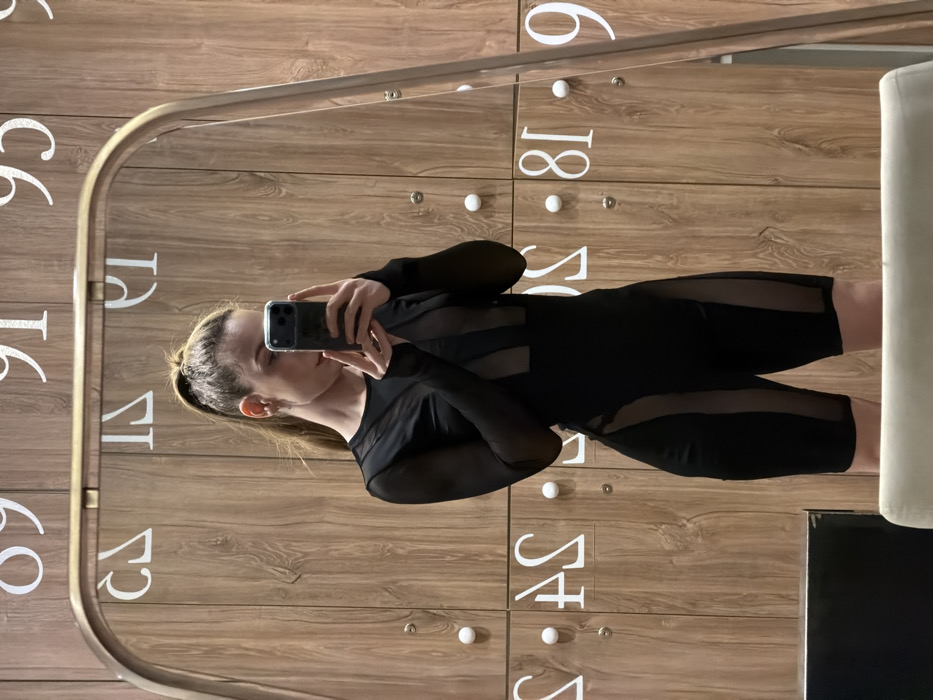 A female individual is depicted capturing her reflection in a mirror, centered within the frame. She wears a black, long-sleeved athletic jumpsuit featuring sheer mesh panels across the sleeves, upper torso, and along the lateral sides of the shorts. Her hair is styled in a high ponytail. She holds a dark-colored smartphone, equipped with a visible triple camera array, in both hands, positioning it to obscure the lower half of her face as she takes the photograph. The background, viewed through the mirror, consists of a wall composed of light wood-grained locker doors, each fitted with a white circular knob and a small keyhole or indent. Prominently displayed on the locker fronts are reversed white numbers and letters, including '89,' '91,' '93,' and '95' in the upper section, and 'AS,' 'S,' '81,' and '9' in the lower visible areas. The mirror itself is partially framed at the top by a thin, rounded, light-metallic border. The scene takes place within a locker room or changing facility. The specific location within Moscow, Russia, cannot be identified from the visual information provided.