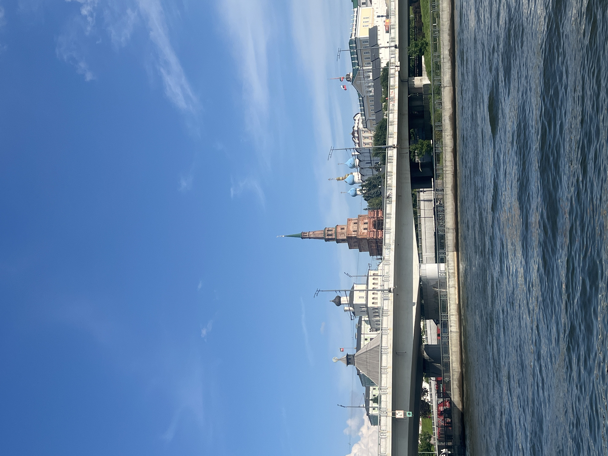 The image shows a view of the Kazan Kremlin from across the Kazanka River. A modern bridge spans the river in the foreground, with a body of blue water below. On the far bank, the historic Kazan Kremlin complex is visible, featuring a tall, multi-tiered brick tower and several domed buildings. The sky is bright blue with scattered white clouds. The scene depicts a cityscape with both modern and historical architectural elements.