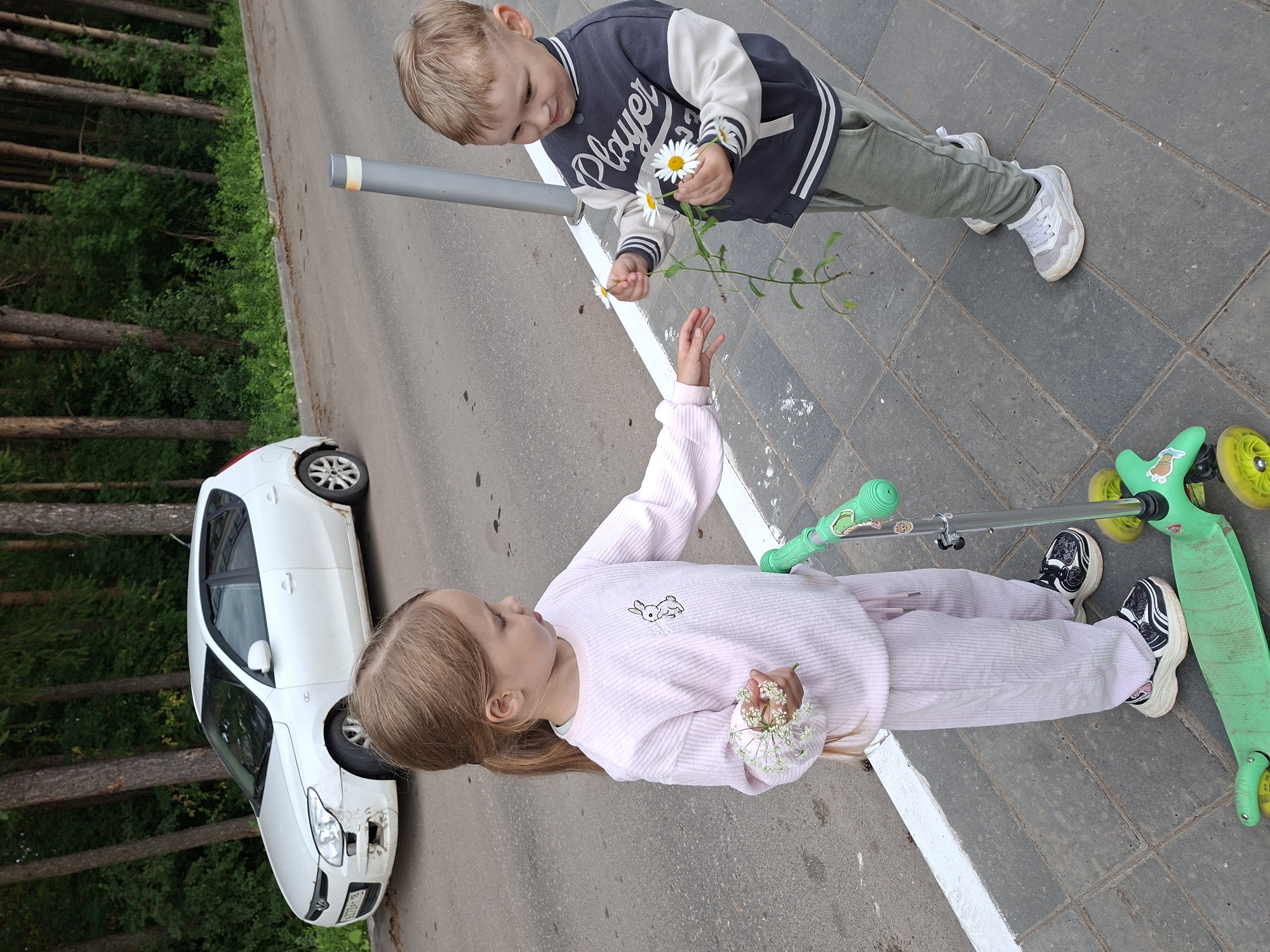 In Izhevsk, Russia, two young children are interacting on a sidewalk next to a paved road bordered by trees. A white hatchback car is parked on the side of the road. The girl, wearing a light pink tracksuit with a rabbit emblem, holds a small bouquet of white flowers. The boy, dressed in a dark gray and cream jacket with "Player" written on it and olive green pants, offers her daisies. A green scooter is positioned in front of the girl. The scene is captured in daylight with natural lighting.