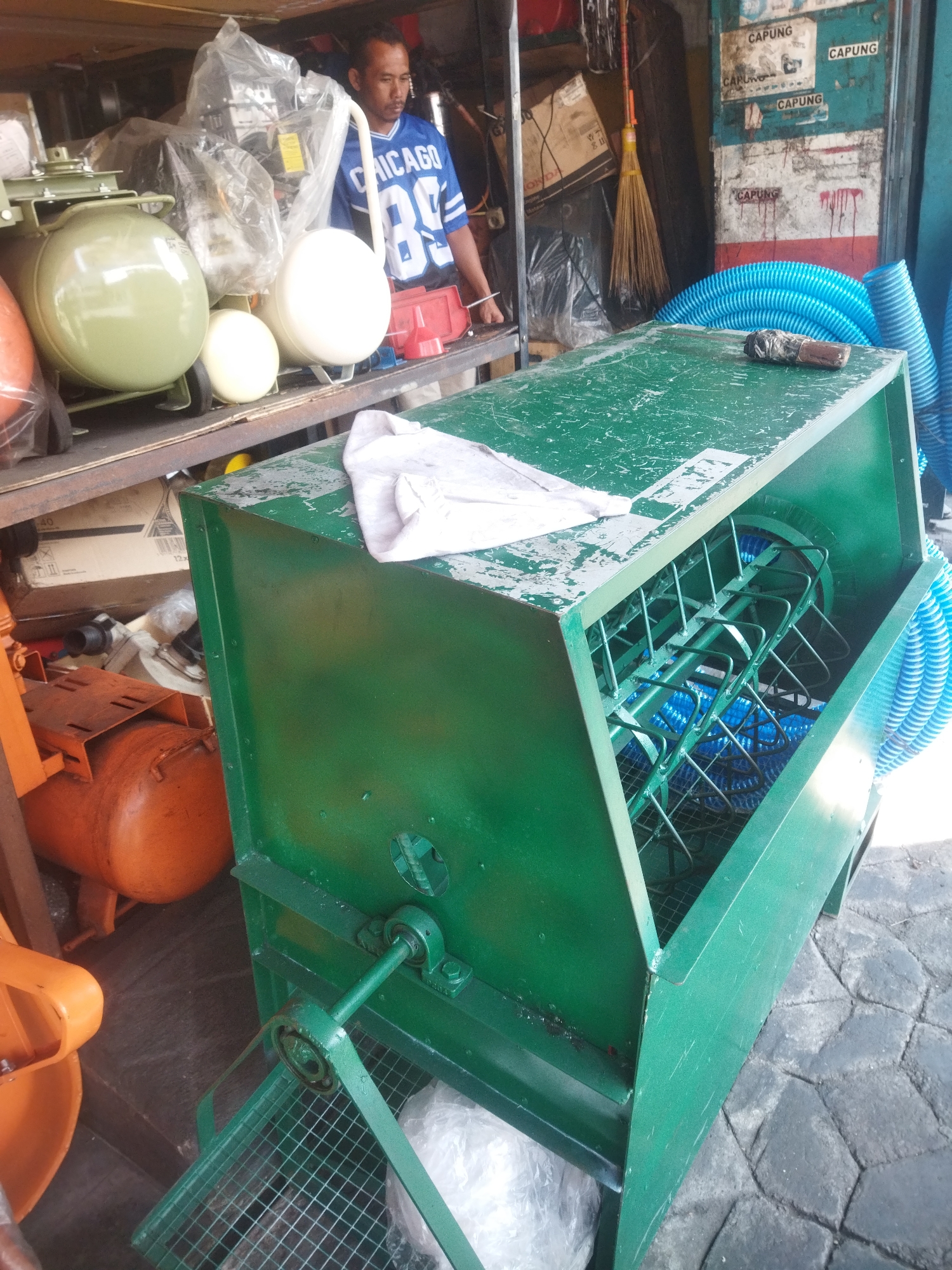 In Giripurwo, Indonesia, a man wearing a blue and white "Chicago" sports jersey stands behind a large green industrial machine. The machine, which appears to be a type of grinder or mixer, has a cage-like interior filled with blue tubing. To the left of the machine, several air compressors are visible, one with a large olive-green tank and another smaller white one. In the background, a broom with a yellow handle leans against a wall covered with faded blue and white posters. The ground is paved with cobblestones.