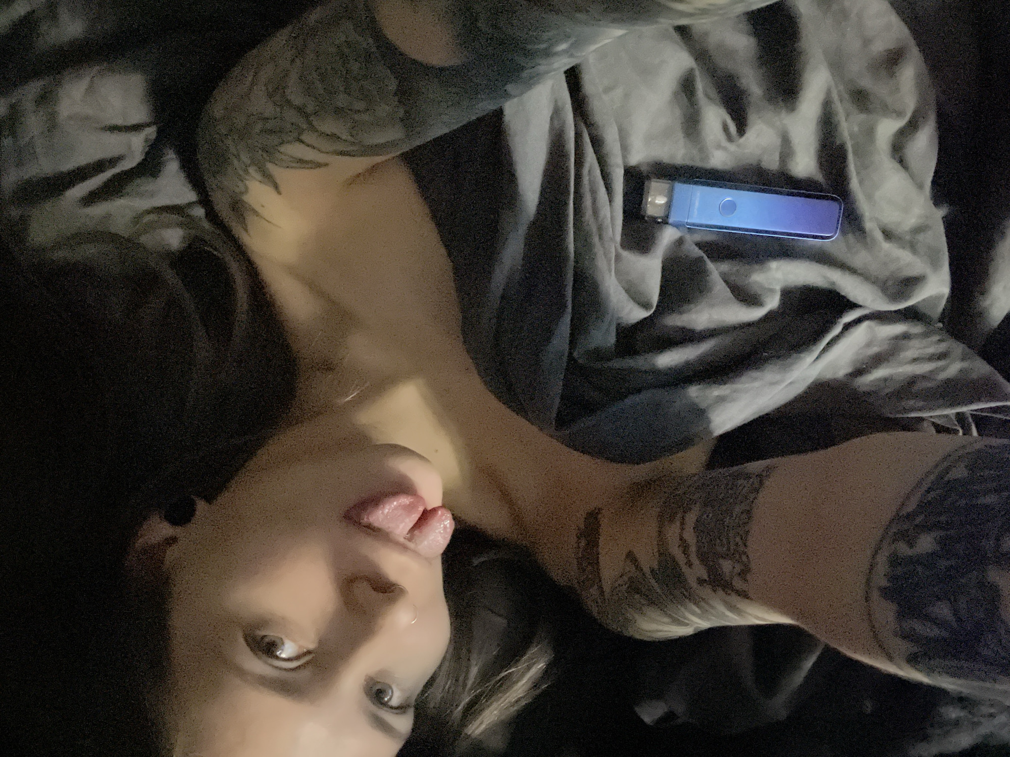 An indoor, dimly lit scene depicts a female subject lying on dark grey bedding. The subject's head is tilted, with light-colored eyes looking directly towards the camera. She has dark hair with lighter highlights. A small ring is visible in her left nostril. Her tongue is extended from her mouth. She appears to be partially uncovered, with her bare shoulders and upper chest visible above the bedding. Both of her visible arms, including shoulders and forearms, are extensively covered with dark, intricate tattoos. A blue and purple gradient vaping device, rectangular in shape with a visible mouthpiece and a circular button, rests on the dark grey bedding to the right of her torso. The environment is obscured beyond the immediate bedding, offering no discernible details about a specific room or location. There are no visual elements within the image to identify the location as Khimki, Russia, or any specific address within it.
