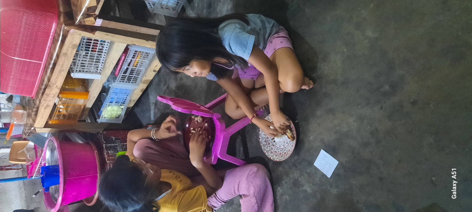 In the city of Jatitengah, Indonesia, two young girls are seated on the floor, engaged in eating. The girl on the left, wearing glasses and a yellow t-shirt, is actively handling food from a plate placed on a small pink chair. She is dressed in pink trousers. The girl on the right, wearing a light blue t-shirt and pink shorts, sits opposite her, also in the process of eating. A patterned plate with food is positioned between them on the floor. The environment appears to be an indoor setting with a concrete floor. In the background, a wooden shelf unit is visible, filled with various plastic containers and baskets. To the left, a large pink basin and a blue container are also present. A white piece of paper with some text is placed on the floor in front of the girl on the left. The image is captured with a "Galaxy A51" phone.
