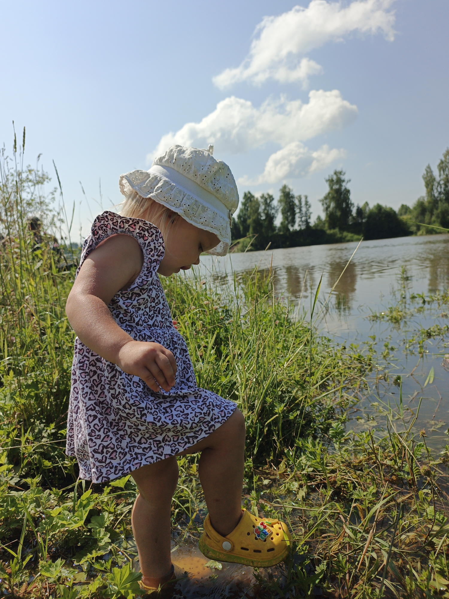 In Urtiushka, Russia, a young girl with blonde hair, wearing a white eyelet sun hat and a white dress with a pink heart pattern, stands on the bank of a body of water. Her right leg is extended, with her yellow Croc shoe submerged in shallow water. She is looking down towards the water and grass. Tall green grass and reeds surround her, with a calm body of water in the background reflecting the sky. The sky is blue with scattered white clouds, and trees line the distant bank.