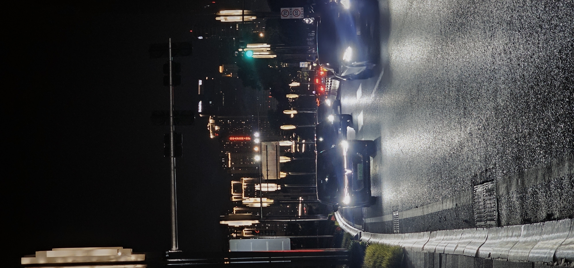 The image depicts a nighttime urban scene in 江北城街道, China. The perspective is oriented to show a street with multiple lanes, and several cars moving along it, their headlights reflecting on the wet pavement. Buildings are visible in the background, with their lights creating a blurred effect. A traffic light in the distance is green, indicating the flow of traffic. The foreground shows part of a concrete barrier and greenery along the side of the road. A street light is also present on the left side. Road signs with speed limits are visible in the distance.