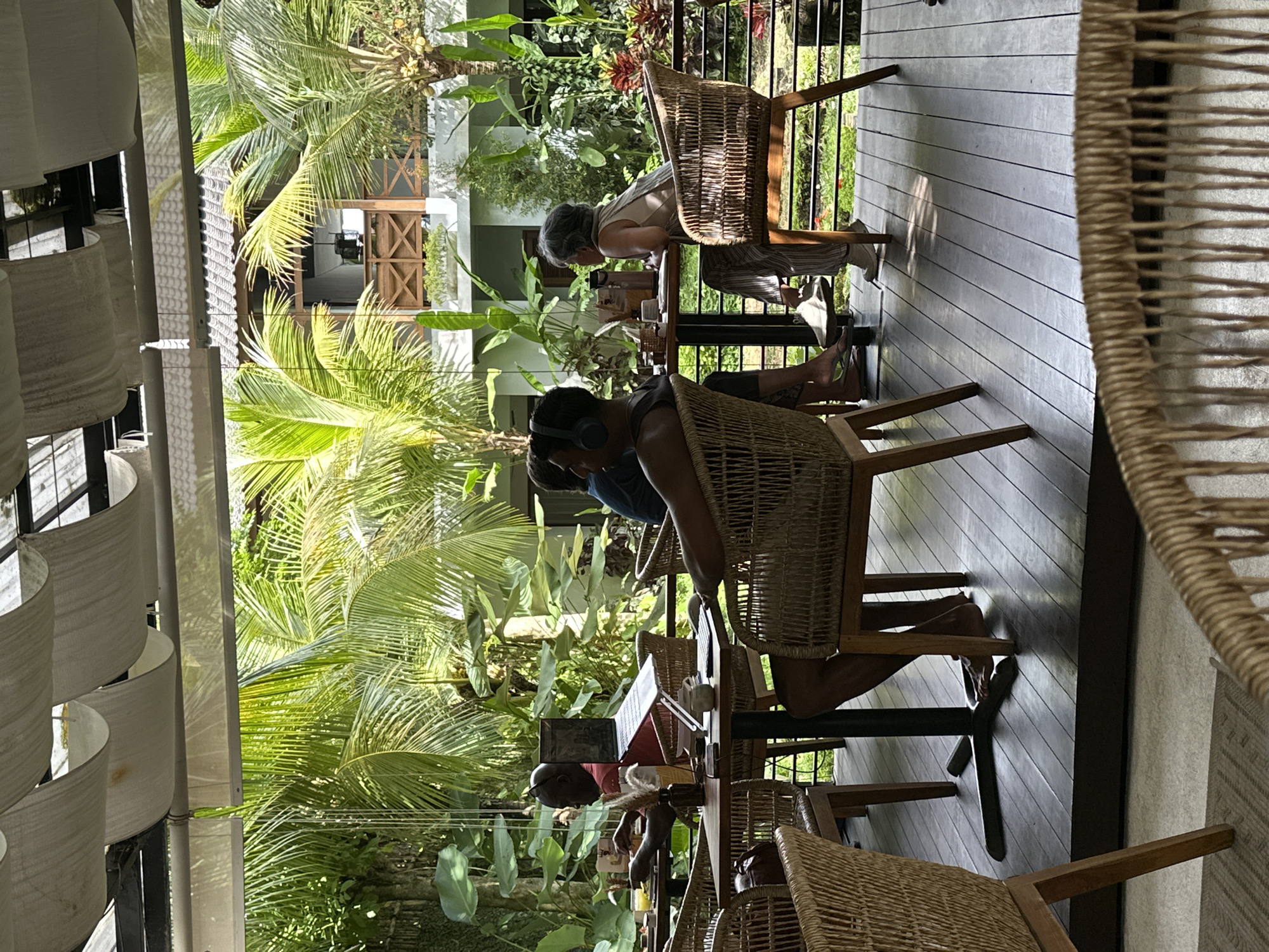 The image shows people sitting at tables on a wooden deck surrounded by lush greenery, including palm trees. The scene appears to be outdoors, possibly a restaurant or cafe in a tropical location. Some people are working on laptops, while others are relaxing. The seating includes wicker chairs. There is an outdoor building in the background.