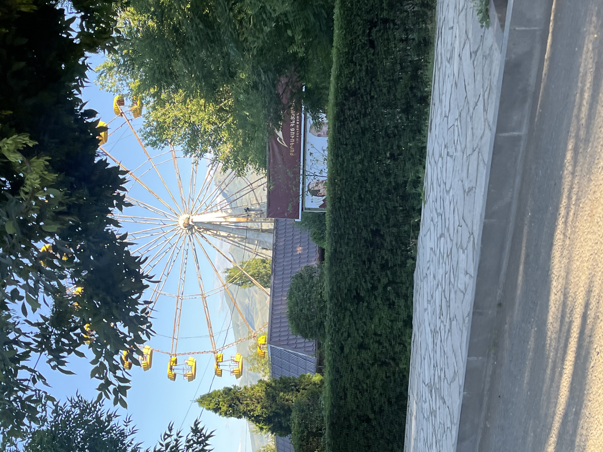 This is a vertical, eye-level shot of a Ferris wheel in an outdoor setting. The Ferris wheel is large and appears to be made of white metal, with numerous spokes radiating from the center. Several yellow gondolas hang from the wheel, some empty and some with the suggestion of people inside. The Ferris wheel is set against a clear blue sky, with the tops of green trees obscuring parts of the wheel.
In the mid-ground, behind the Ferris wheel, is a building with a dark, tiled roof. A banner is visible hanging between two trees, displaying some text in Armenian script. Two portraits are also visible on the banner, one of a man in a suit and the other of a man with glasses.
To the right of the Ferris wheel, there is a tall, neatly trimmed hedge. Beyond the hedge, a wall constructed of light-colored irregular stones is visible. The lighting suggests it is daytime, possibly late afternoon due to the shadows cast on the stone wall. The overall scene appears to be a park or amusement area on a sunny day.