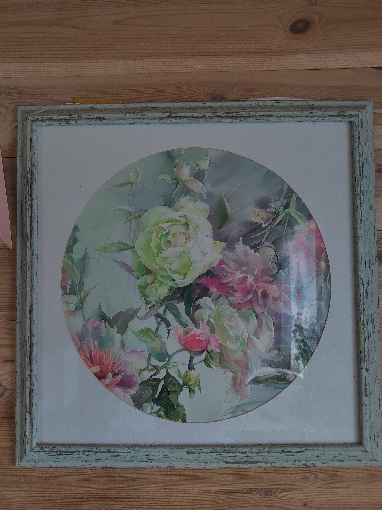 The image shows a framed circular watercolor painting of peonies, displayed on a wooden wall. The painting features a variety of peony blossoms in shades of light green, pink, and white, with dark green leaves and some buds. The frame is light blue-green with a distressed, chippy paint finish. The wooden wall behind the painting has visible grain and knots. The scene appears to be indoors. There is no discernible location in the city of Бярэзінскі сельскі Савет, Belarus.