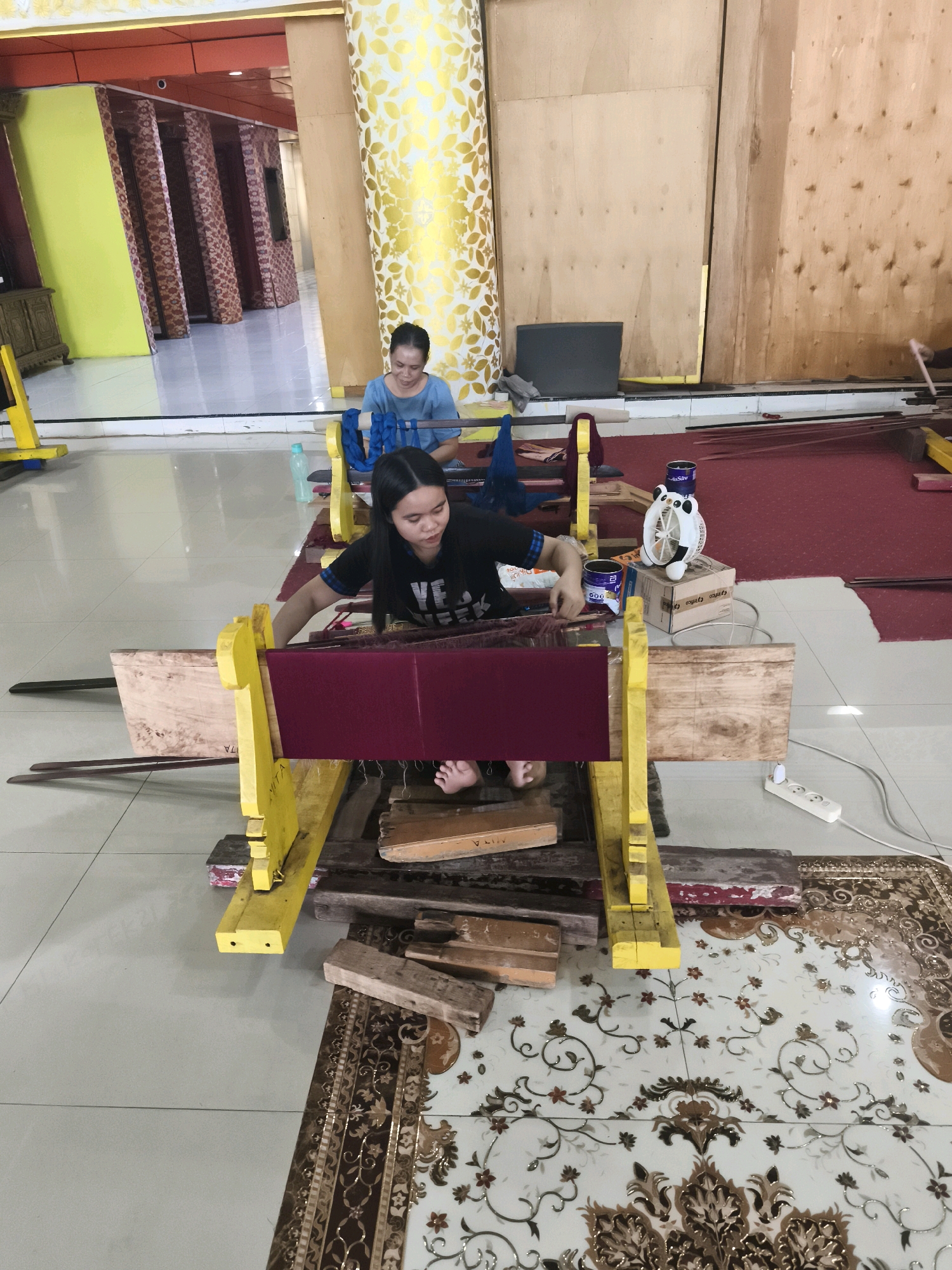 The image captures a scene of textile production in what appears to be an indoor workshop or studio in Palembang, Indonesia. In the foreground, a young woman with long dark hair is actively engaged in weaving on a loom. She is wearing a black t-shirt with white lettering. Her hands are positioned on the loom, and her feet are visible, suggesting she is operating the loom by foot pedals or a similar mechanism. The loom itself is constructed from wood and painted bright yellow. A piece of deep red fabric is stretched on the loom, indicating the current weaving project.
Behind the woman, another woman is seated and appears to be observing or assisting. She is wearing a blue t-shirt. The background reveals a spacious room with tiled flooring and wooden walls. Several pillars decorated with gold leaf patterns are visible, adding an element of traditional Indonesian architecture. There are also some rolls of fabric, wooden planks, and a small, cartoon-shaped fan on a box, all suggestive of an active workshop environment. The overall impression is one of a traditional craft being practiced in a functional, if somewhat rustic, setting.