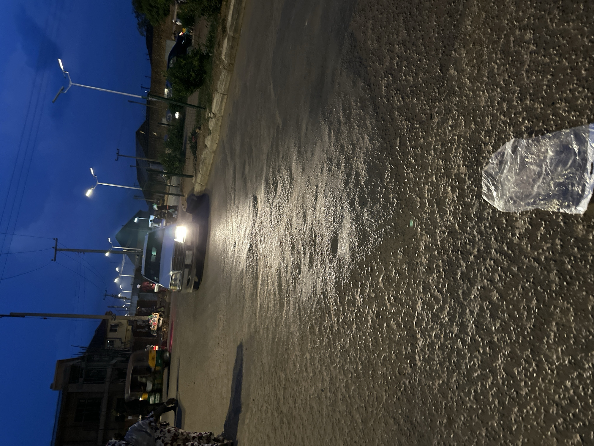 The image depicts a street scene at dusk in Bukuru, Nigeria. The sky is a deep twilight blue, with visible streetlights illuminating the paved road. In the foreground, the asphalt appears wet and reflective, with scattered small, light-colored particles creating a textured surface. A crumpled, transparent plastic bag lies on the road in the lower right quadrant of the image.
Further down the street, a silver car with its headlights on is approaching. To the left of the car, a three-wheeled auto-rickshaw, commonly known as a "Keke Napep," is visible. A few figures, possibly pedestrians, are present near the vehicles and buildings lining the street. One person on the far left is wearing a patterned garment. Buildings, some appearing under construction, are situated along the left side of the road. Various street vendors' stalls with merchandise are also apparent near the intersection. The environment suggests a typical urban street scene in Nigeria during the evening transition from day to night.