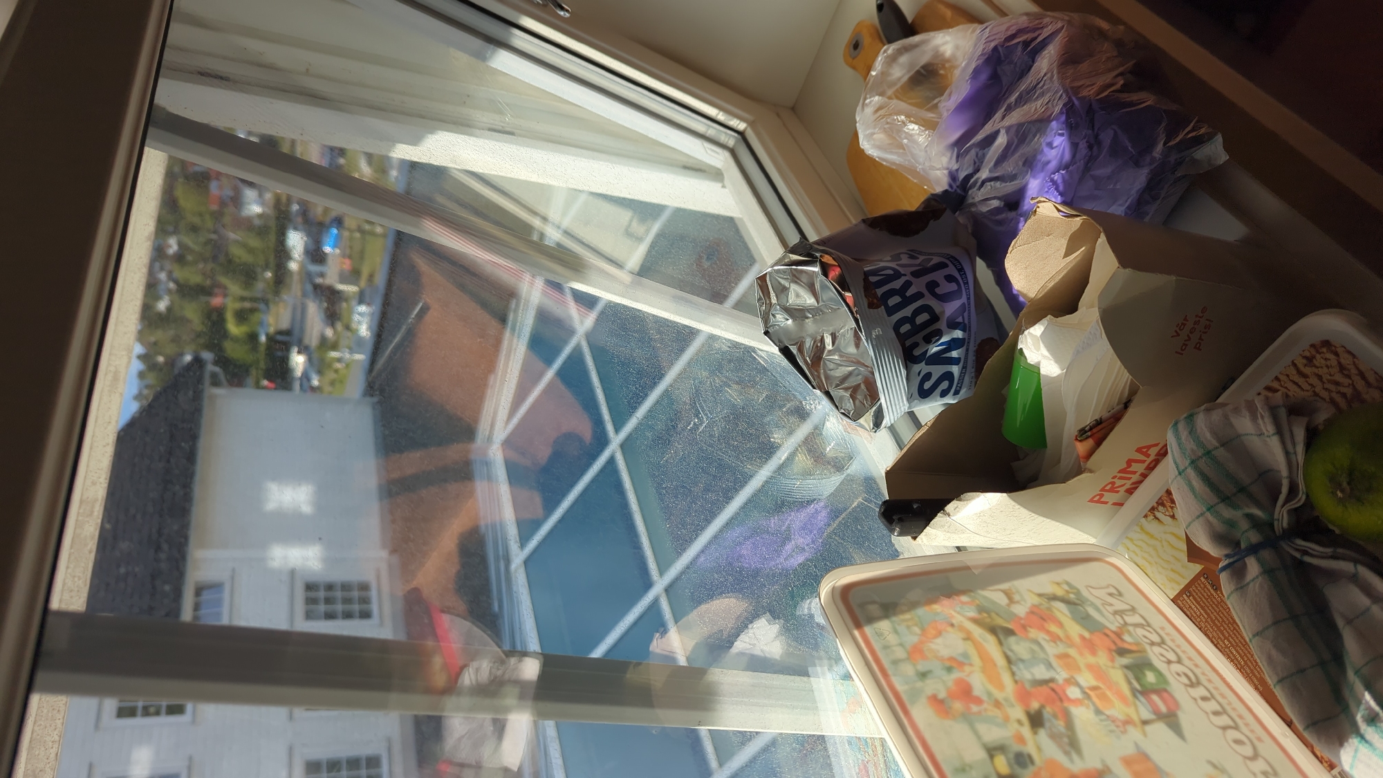 The image shows a close-up view of a windowsill in Larkollen, Norway, with a collection of items on it. Through the window, a white building with a dark roof is visible, situated in a residential area with trees and other houses in the background. The window itself is grimy, with reflections of the interior on the glass.
On the windowsill, from left to right, is a rectangular tray with an illustration of people in orange outfits. Next to it is a cardboard box containing various items, including white paper napkins, a green cup, and a bag of "SABRO SNACKS". To the right of the box is a clear plastic bag filled with purple items, possibly clothing or fabric. A checkered cloth is laid out in front of the box, with a green lime in the bottom left corner. The overall impression is of a slightly cluttered but lived-in space, likely a kitchen or dining area.