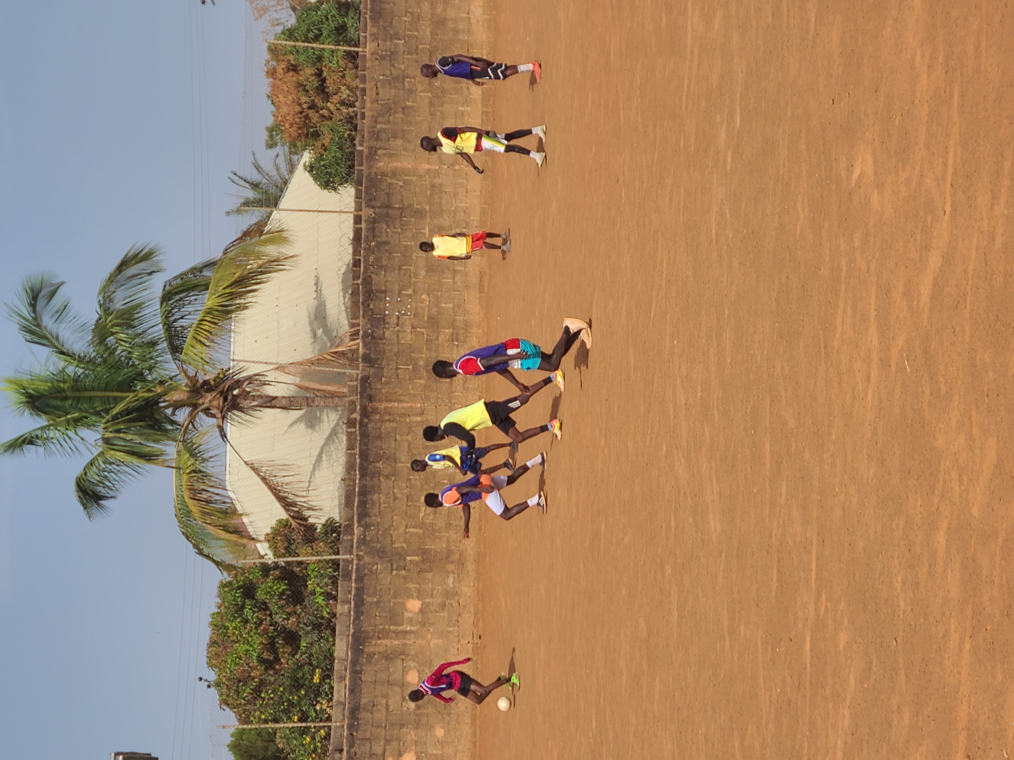 A sunny outdoor scene in Barakin Lerawi, Nigeria, shows a dirt field with a brick wall along one side. Seven young men in various athletic wear are spaced along the length of the wall, appearing to be performing a physical exercise, likely stretches or warm-ups. One young man in the foreground is kicking a soccer ball. In the background are trees, a building, and a palm tree, suggesting a residential or recreational area.