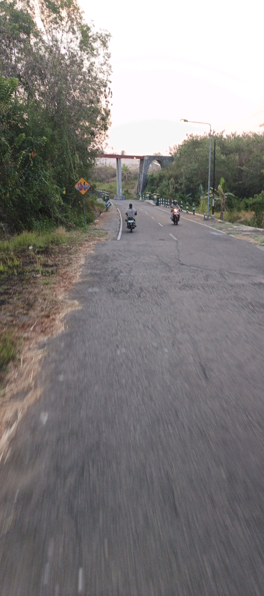 In Sumberrahayu, Indonesia, a two-lane asphalt road curves through a rural landscape under a pale sky, likely at dusk or dawn. The road surface shows some wear and tear, with faint white lines marking the lanes. On the left side of the road, dense green foliage and dry grass line the shoulder. A diamond-shaped warning sign with a yellow background and a red border, depicting what appear to be stylized figures, stands prominently on the left.
Further down the road, a concrete bridge with a red steel superstructure arches over the pathway, suggesting it's part of a larger infrastructure project. Along the sides of the road, there are short posts with reflective white and black barriers.
Several individuals are on motorcycles, traveling in the same direction as the camera. One motorcycle, ridden by a person wearing a grey shirt, is in the left lane. Another motorcycle, with a rider wearing a red shirt, is in the right lane. The motorcycles are spaced apart, indicating individual travel. A third motorcycle is visible further ahead in the right lane. A lamppost stands on the right side of the road, near the bridge. The surrounding environment is lush with trees and vegetation. The perspective suggests the image was taken from a moving vehicle, likely a motorcycle, due to the motion blur and the low angle.