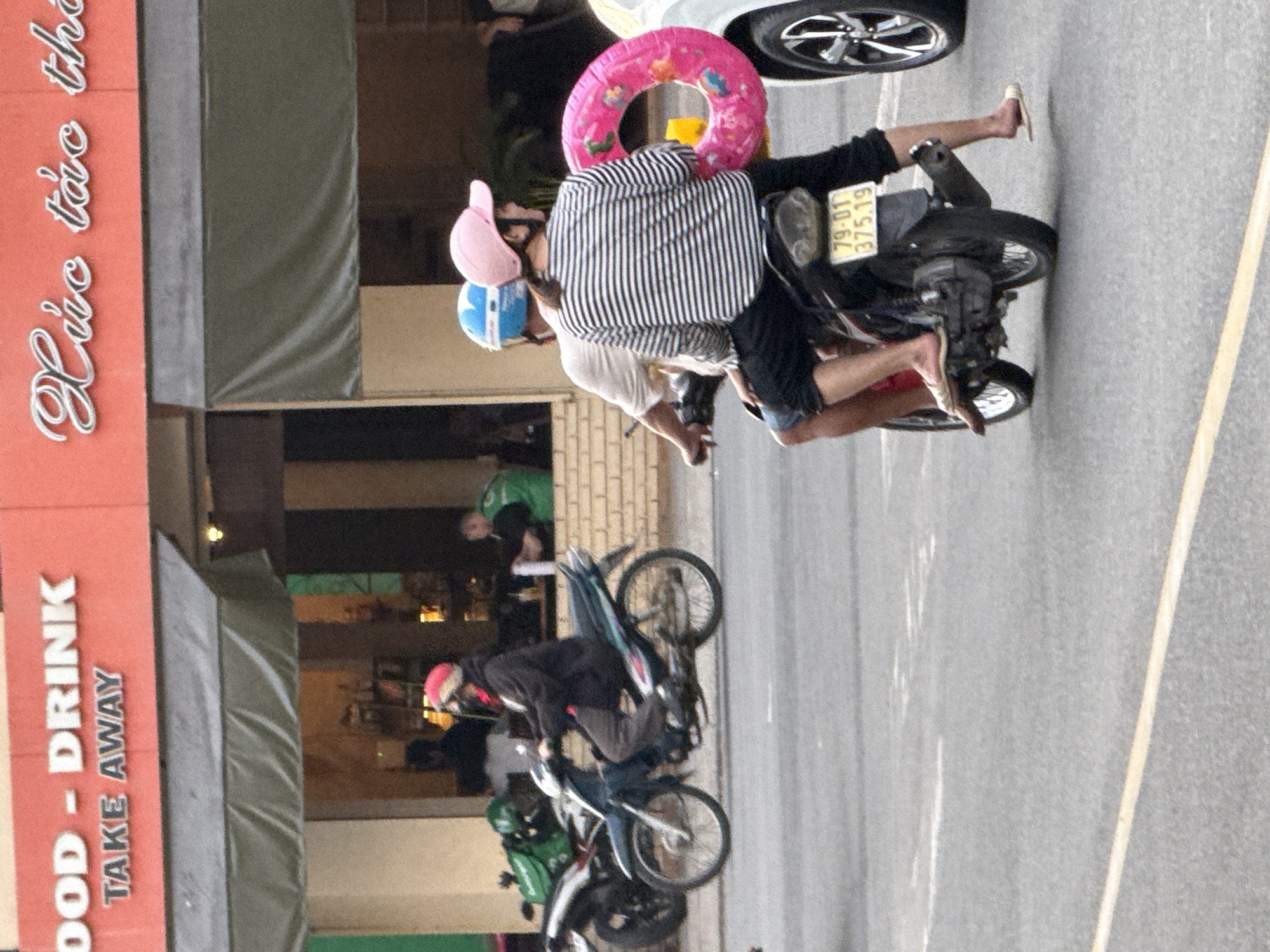 This is a street-level, medium shot captured in Nha Trang, Vietnam, likely during the daytime. The scene features two individuals riding a motorbike. The person in the front, wearing a blue helmet, is driving. The person in the back, wearing a pink baseball cap and a striped long-sleeved shirt, is holding a large pink inflatable ring. Both are wearing casual attire.
The motorbike has a visible license plate with the number "79-D1 375.19". To the left of the motorbike, another motorbike with a green storage box is parked. In the background, a building with a red facade is visible, displaying signage that reads "FOOD - DRINK" and "TAKE AWAY" in English on the left side. The right side of the building has Vietnamese text written in a cursive font. An awning extends from the building over the street. Further in the background, people are visible inside what appears to be a food establishment. The pavement is asphalt, with a yellow painted line indicating a road boundary. A white car is partially visible on the right side of the image.