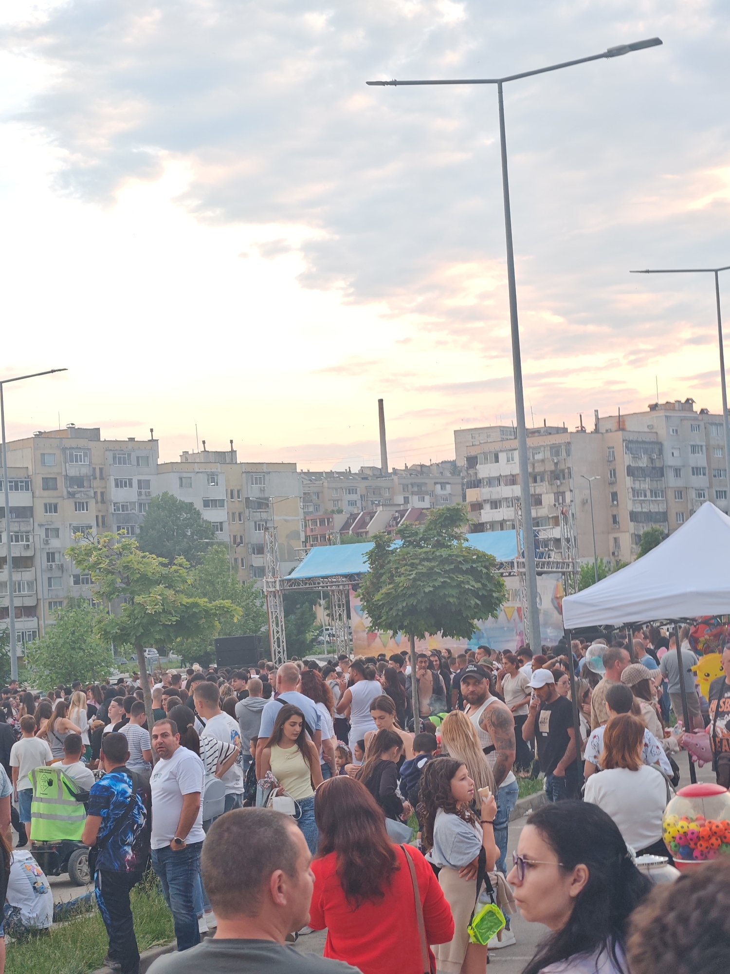 The image shows a large crowd of people gathered in an open area. In the background are residential buildings. There is a stage structure with blue tarp covering the top. There is a white tent. The image shows several street lamps. The gathering appears to be an event or festival. The location seems to be in Shumen, Bulgaria, as implied by the prompt. The overall tone is one of a community gathering. The people are diverse in appearance, including men, women and children of various ages and ethnicities.