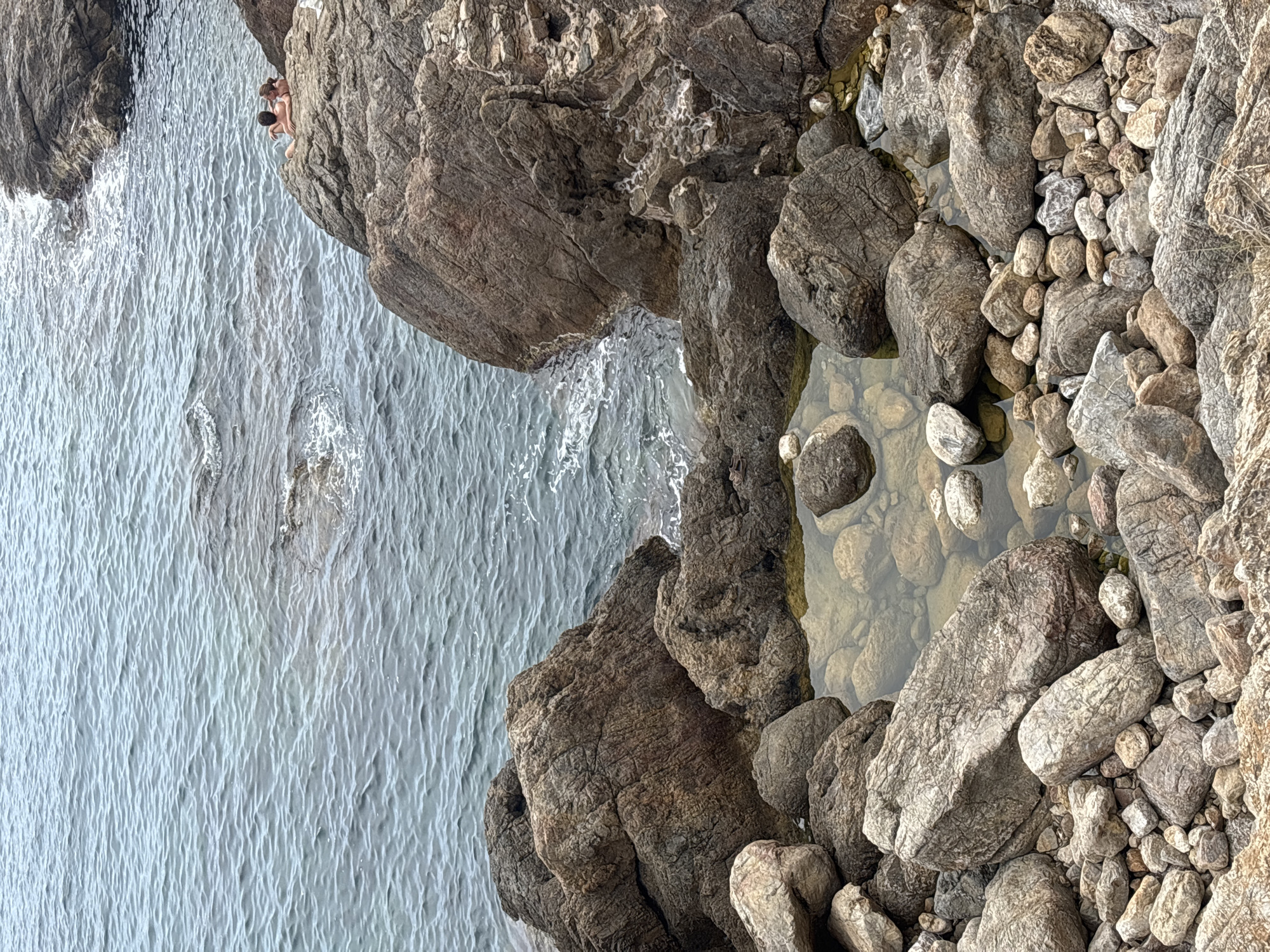 The image shows a rocky coastline with calm, blue water. In the upper right portion of the image, two people are sitting on a large rock overlooking the sea. In the foreground, a tide pool is formed amongst smaller rocks, with clear water revealing the pebbles beneath. The rocks are various shades of brown and grey, with some showing signs of weathering and erosion. The scene appears to be a natural coastal landscape, likely in a Mediterranean climate, consistent with locations such as Begur, Spain.