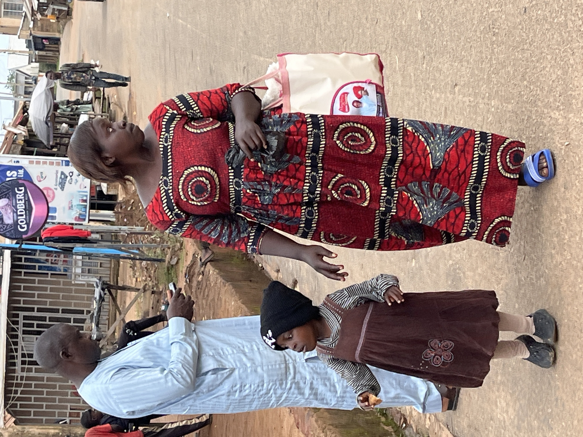 A man wearing a pale blue dashiki stands facing right, looking down at a mobile phone in his hands. Beside him, a young child in a brown dress, striped shirt, and black beanie is looking to their left. To their right, a woman in a vibrant red and black patterned dress is looking to her left, carrying a white tote bag in her right hand. The scene appears to be a street in Jos, Nigeria, with buildings and market stalls visible in the background.