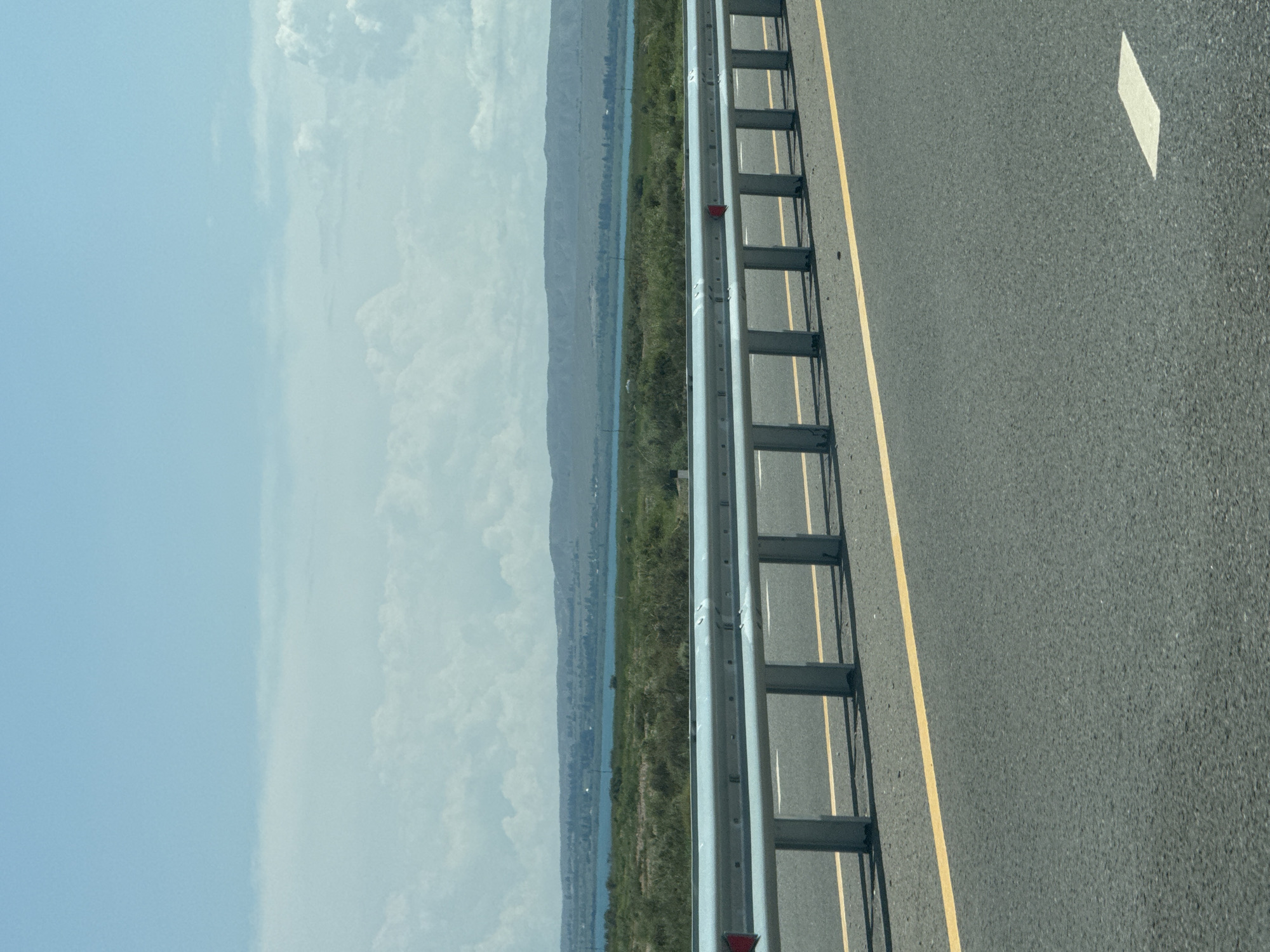 The image depicts a highway scene in Balykchy, Kyrgyzstan, on a clear day. In the foreground, a section of asphalt road is visible with a yellow lane line and a white lane marker. A metal guardrail runs along the side of the road, with several vertical supports and reflective red markers. Beyond the guardrail, there is a strip of green vegetation, and further in the distance, a body of water, likely Lake Issyk-Kul, can be seen. A range of rolling, barren hills forms the background, under a sky with scattered white clouds. No people or vehicles are visible in the image.