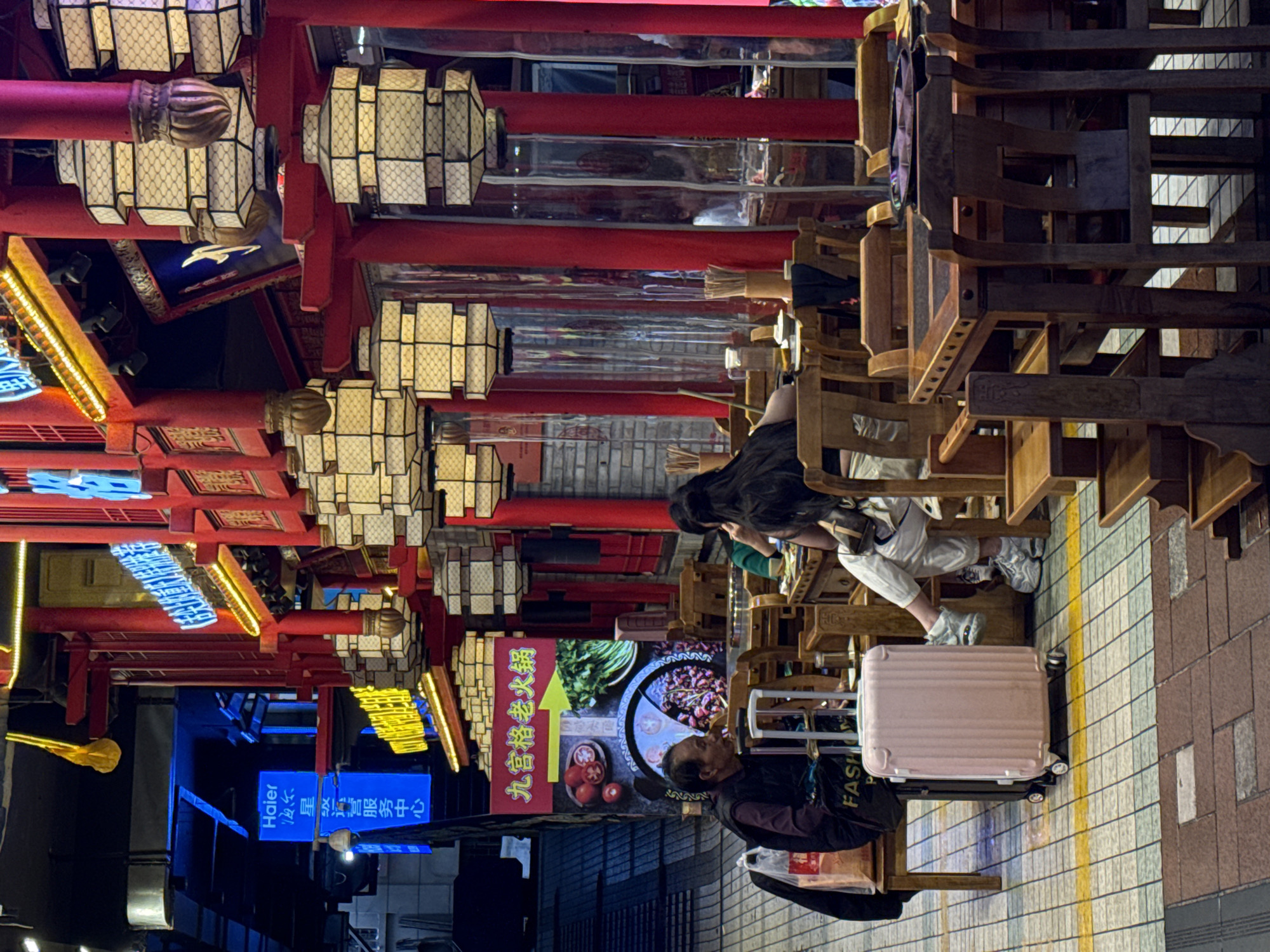 The image depicts an outdoor dining area in Yuzhong District, China. The scene features a bustling environment with people seated at wooden tables and chairs. Lanterns and neon signs adorn the overhead structures, adding to the atmosphere. A pink suitcase is visible near one of the tables, suggesting a traveler is present. The focus is on the dining experience and the urban ambiance.