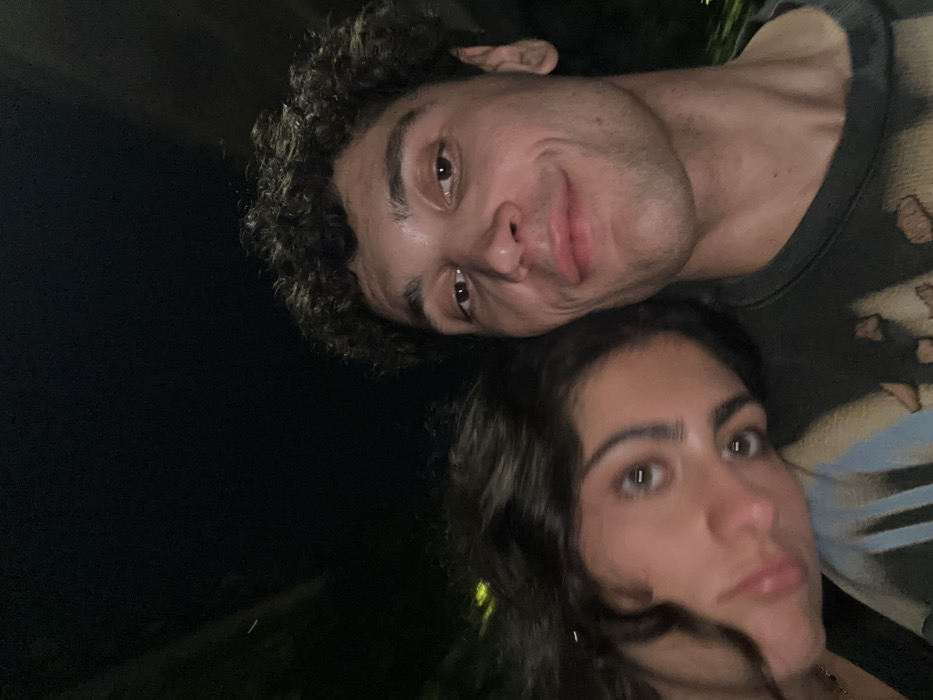 Two people pose closely for a selfie. A man with curly hair and a slight smile is positioned above and to the right. Below him is a woman with wavy dark hair and a neutral expression. The background is dark, suggesting nighttime or an indoor setting with dim lighting. Parts of the man's printed t-shirt are visible. Their faces are the focal point of the image.