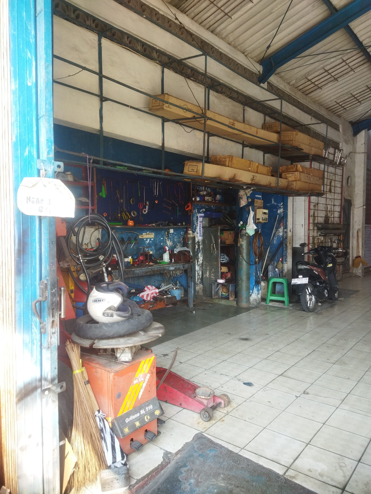 The image depicts the interior of a motorcycle repair shop located in Wonogiri, Indonesia. The shop is filled with various tools and equipment associated with motorcycle maintenance. In the foreground, a tire-changing machine is visible, with a motorcycle helmet and a tire placed on top of it. A broom leans against the machine. The walls are adorned with an array of tools, including wrenches, pliers, and screwdrivers, neatly organized on a pegboard. Shelves above the workbench are stacked with cardboard boxes. In the background, a dark-colored motorcycle is parked. The floor is tiled with white squares. A green plastic stool sits near a large green gas cylinder. The overall impression is of a functional and organized workshop.