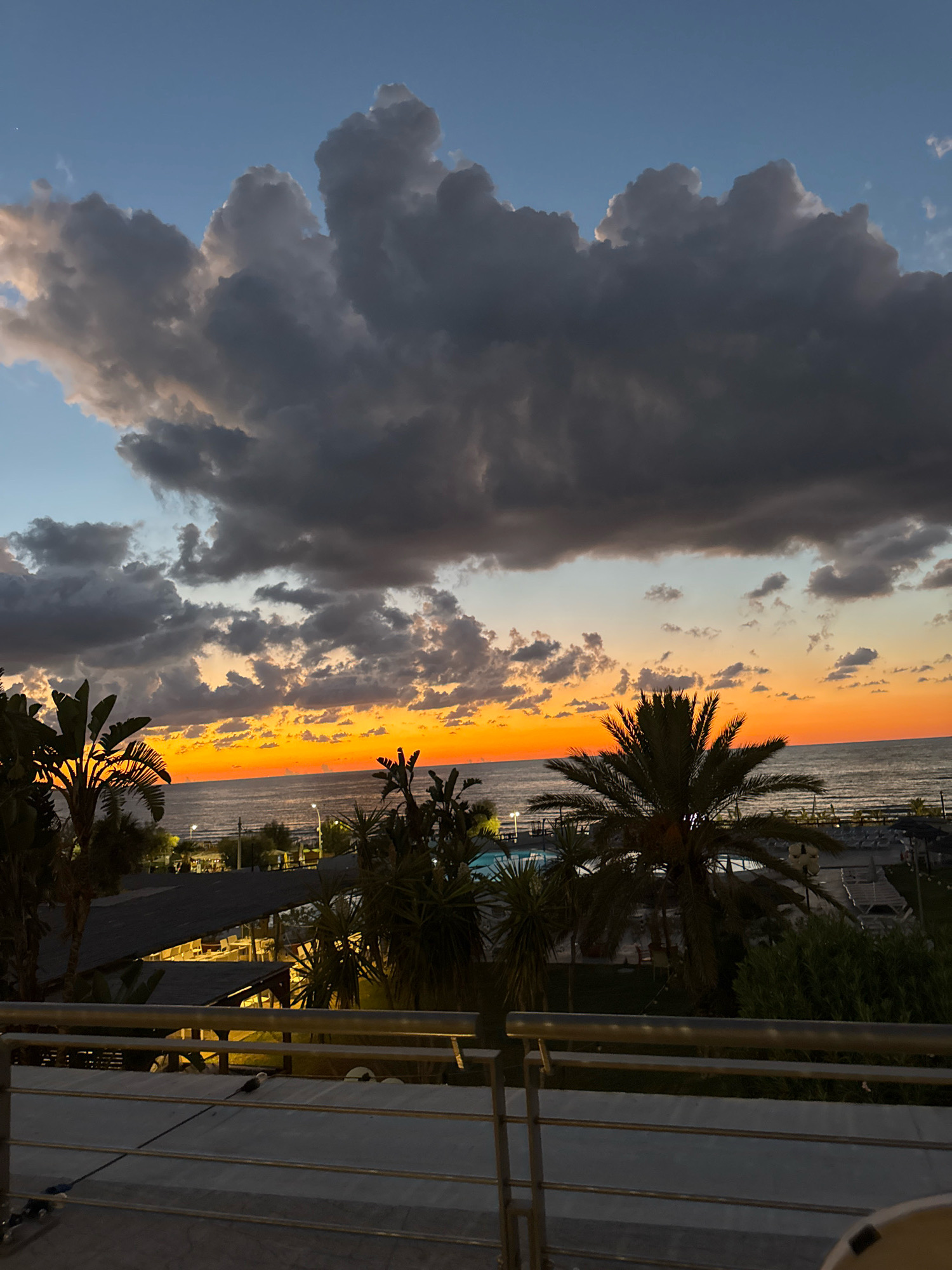 A serene twilight scene unfolds over the sea in Isola delle Femmine, Italy, as the sun dips below the horizon, casting a warm, orange glow across the water and sky. The sky is a canvas of dramatic, dark grey clouds, tinged with the soft light of sunset, juxtaposed against patches of clear blue. Below the vibrant sky, palm trees and other lush greenery frame the view of the coastline.
A resort or hotel is visible in the mid-ground, with structures featuring dark roofs and illuminated interiors, suggesting evening activities. A swimming pool, partially visible, adds to the resort atmosphere. Lounge chairs are arranged on the grass, hinting at a relaxing setting. The sea stretches out to the horizon, calm and reflecting the colors of the sky. In the foreground, a balcony railing made of metal and white material overlooks the scene, suggesting the vantage point from a hotel room or terrace. The overall impression is one of peaceful, late-day tranquility.