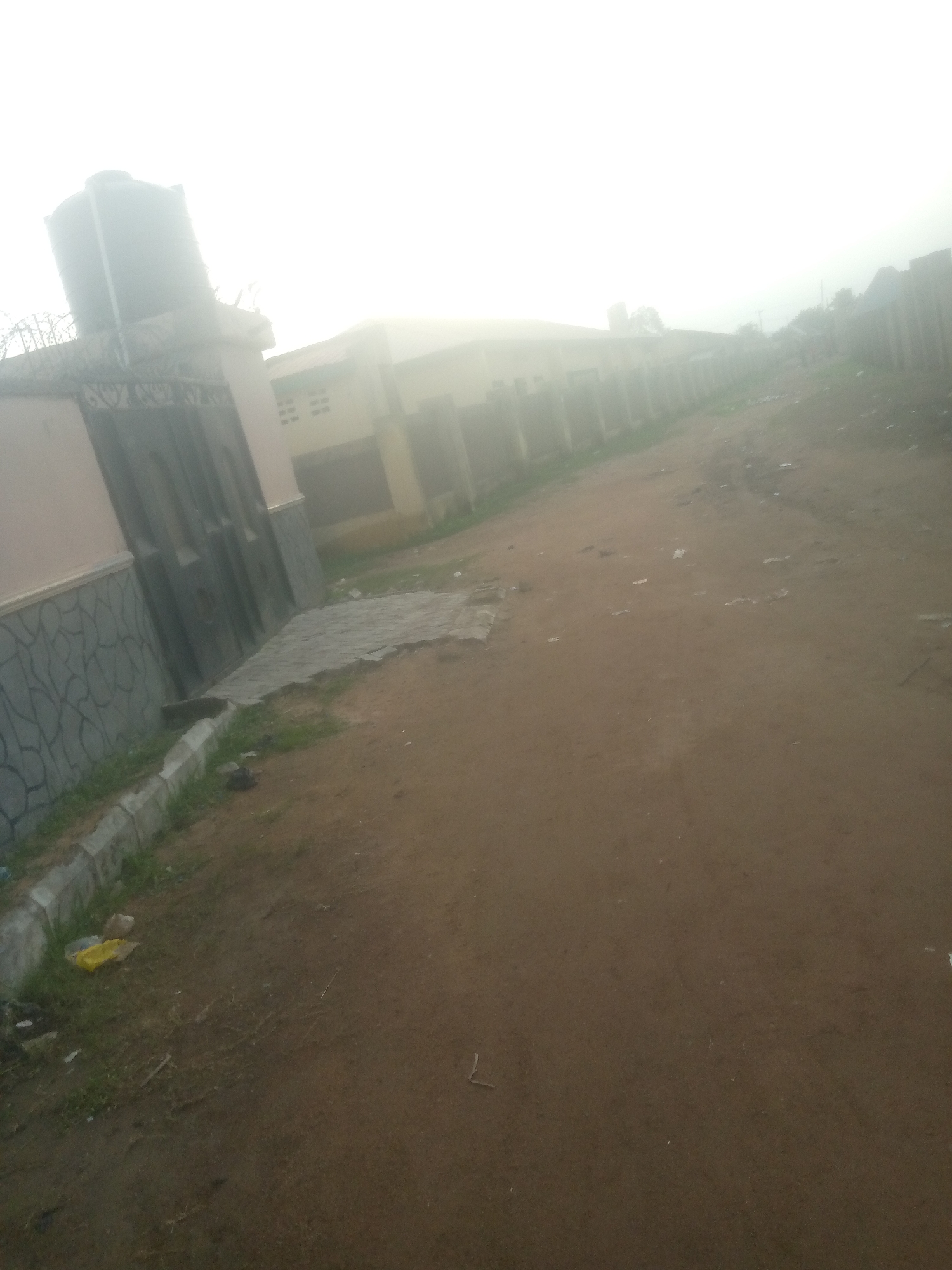 The image depicts a street scene in Palade, Nigeria. The foreground is a dirt road with scattered debris. To the left, a paved walkway leads to a gate with an ornate design. A tall black water tank is visible on top of a building behind the gate. The surrounding buildings are low-rise, with some appearing weathered. The sky is bright and hazy, suggesting overcast conditions or early morning light. The overall atmosphere is one of a modest urban or semi-urban environment.