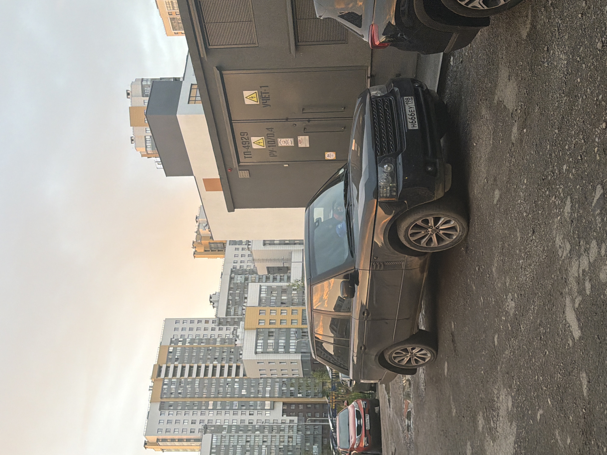 A black Range Rover is parked in a dirt lot in front of a utility building. The Range Rover has a license plate that reads "H666EY 198". A red Kia Sportage is parked to the left of the Range Rover. In the background, there are several tall apartment buildings. The sky is cloudy and the sun is setting, casting a warm glow on the scene. The utility building has two large doors and several signs with Cyrillic lettering and electrical hazard symbols.