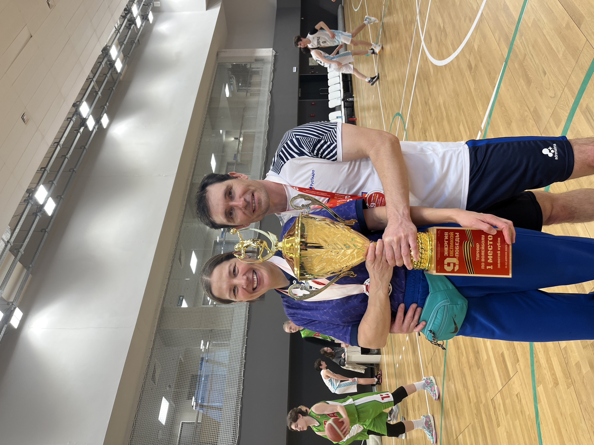 The image shows two individuals standing indoors, presumably in a sports arena, holding a golden trophy and wearing medals around their necks. They appear to be celebrating a victory. The background reveals a basketball court with other people involved in sports activities. The trophy itself has a red placard with text written in Russian, which indicates "1 место" (1st place) and mentions a volleyball tournament held on April 11-12, 2026. The environment suggests a sports-related event or competition, and the people in the foreground seem to be the winners. Additionally, sports apparel is visible on the individuals and in the background. Based on the logo on the shorts, it seems they may be involved with the "adrenalina" brand. Other brands such as "Pusropo" (spelling may be slightly off due to image quality) are also visible on the man's shirt.
