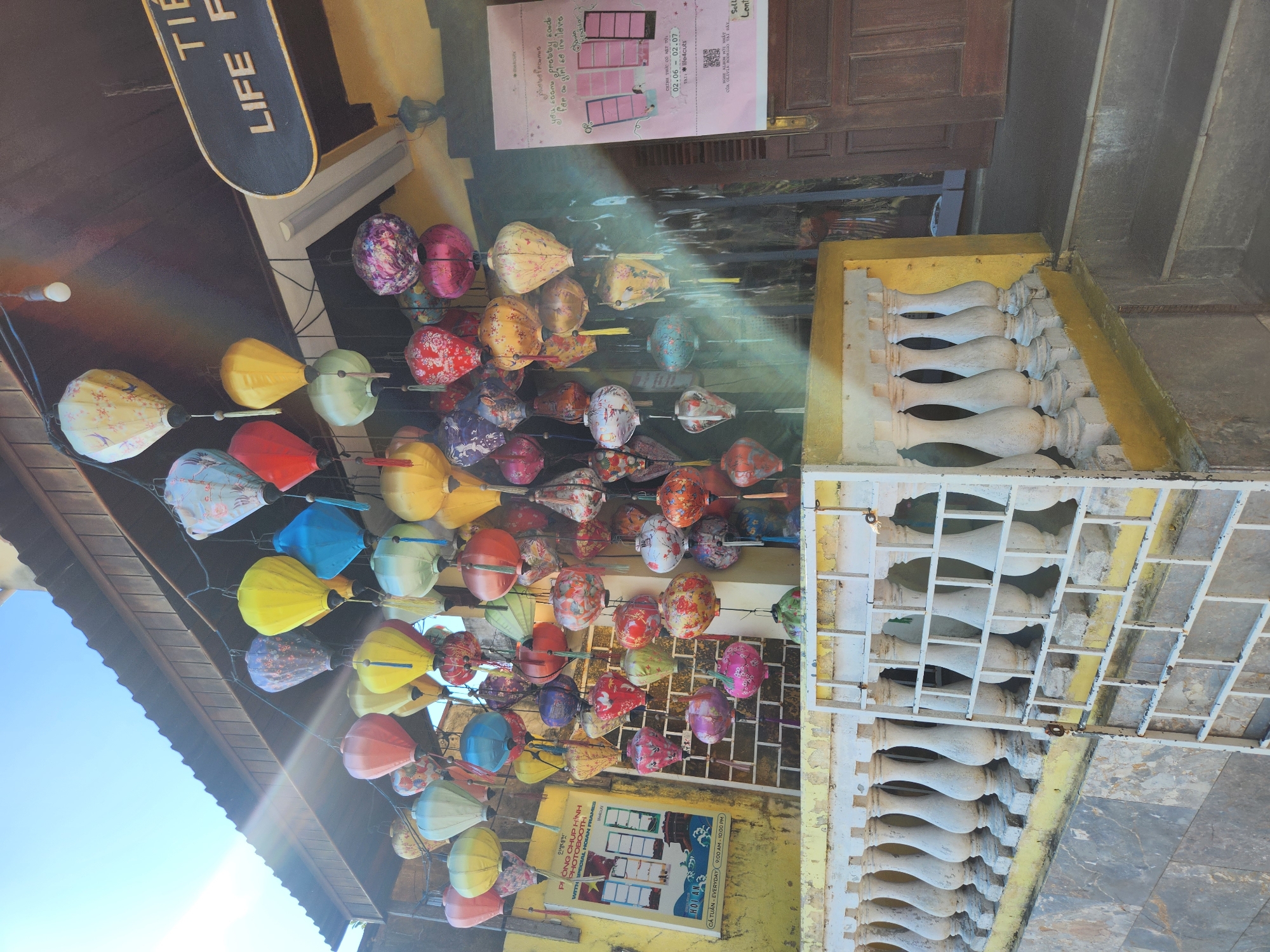 The image shows a colorful display of traditional Vietnamese lanterns hanging from the eaves of a building in Hội An Ward, Vietnam. The lanterns are of various shapes, sizes, and colors, including red, yellow, blue, green, and patterned designs. They are suspended from strings and appear to be made of silk or paper. The building itself has a wooden roof and yellow walls. A white balustrade runs along the front of the building, with a white metal gate or screen leaning against it. A sign with Vietnamese text is visible on the left side of the building, and another sign with "LIFE P..." is visible on the right. The scene is illuminated by sunlight, with lens flare visible in the upper left corner. The overall impression is one of a vibrant and festive atmosphere, typical of the tourist areas in Hội An.