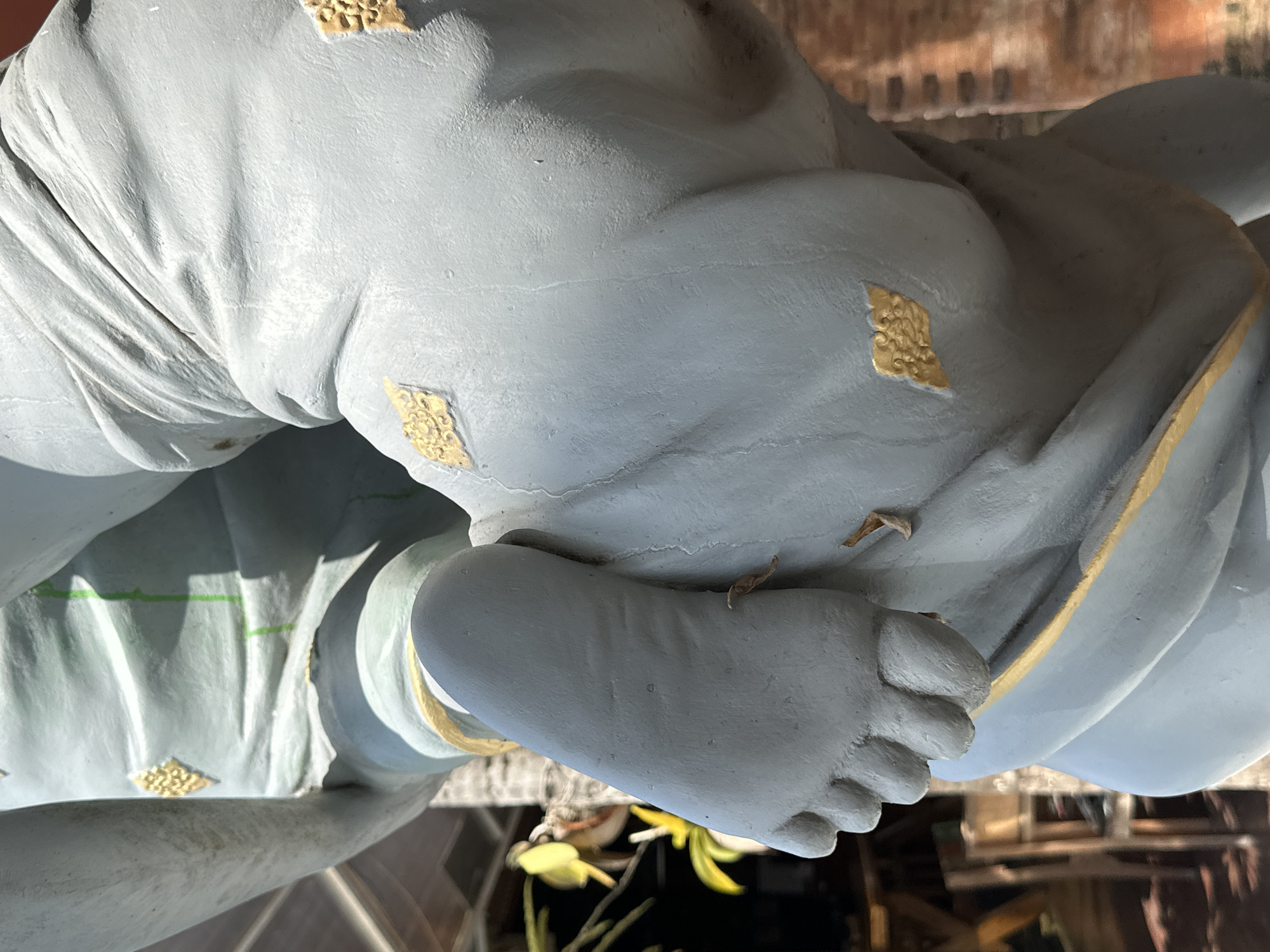 This is a close-up, vertically oriented photograph of a light blue statue, likely made of plaster or concrete, adorned with decorative gold accents. The statue depicts a portion of a human figure, specifically a foot and parts of the torso and leg, which are partially covered by flowing fabric-like material. The gold accents are small, diamond-shaped, and intricately patterned, appearing on the fabric.
The statue is positioned outdoors, as evidenced by the natural lighting and the background which suggests a setting with wooden structures and foliage. The lighting indicates it is daytime, with sunlight casting shadows that highlight the folds and textures of the statue's surface. There are a few small, dry leaves or debris on the statue, suggesting it is exposed to the elements. The overall tone of the image is neutral, with the cool blue of the statue contrasting with the warm tones of the background. No text is visible in the image.