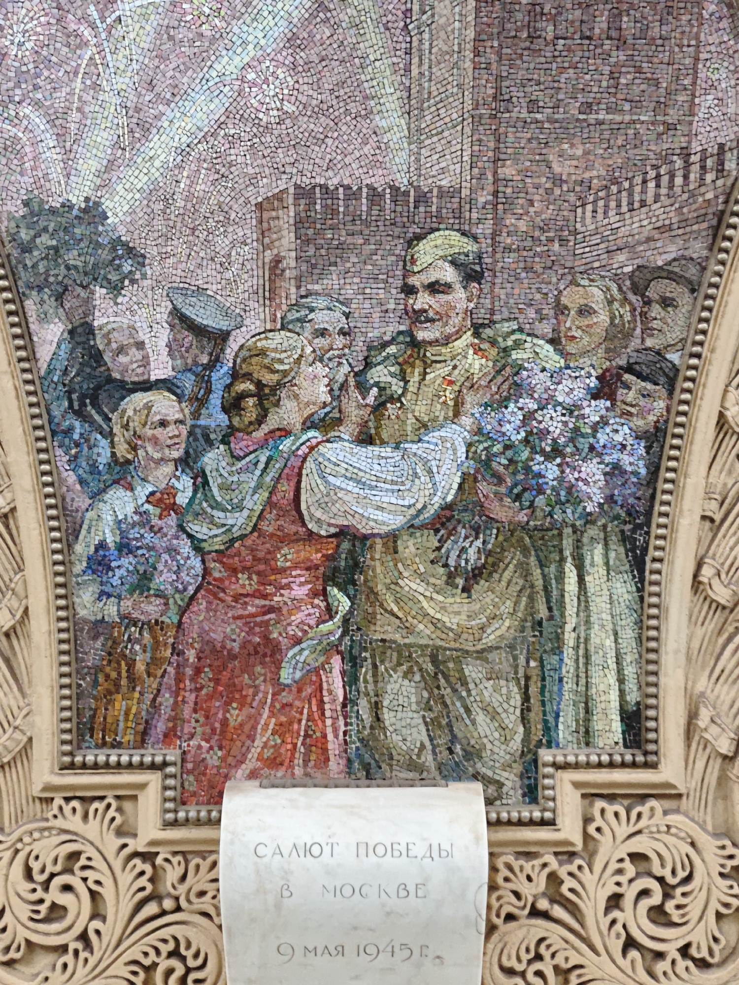 This is a mosaic located in the Kievskaya metro station in Moscow, Russia. The mosaic depicts a celebratory scene with a group of people, likely celebrating Victory Day. In the center, a male figure in military uniform is receiving a bouquet of flowers from a woman. He is adorned with medals. Other individuals in the crowd are also holding flowers, and some are looking on with smiles. In the background, there appears to be fireworks illuminating the sky, and a brick building is visible. Below the mosaic is a stone inscription that reads "SALUTE OF VICTORY IN MOSCOW 9 MAY 1945". The mosaic is framed by ornate golden detailing.