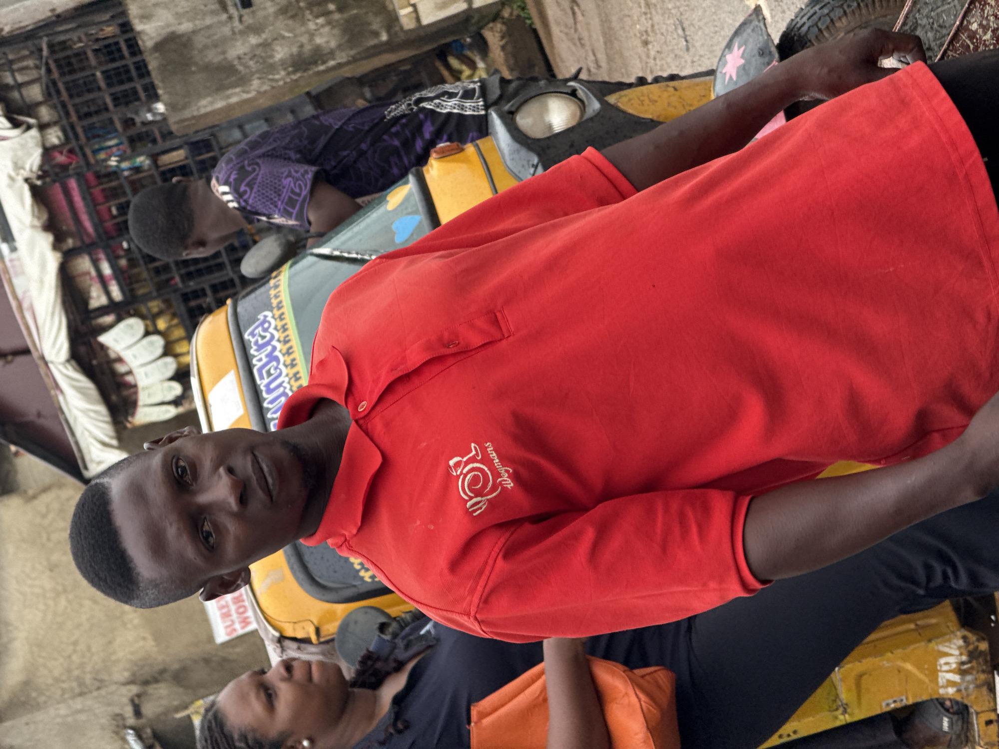 In Abuja, Nigeria, a man in a bright red polo shirt, featuring a white logo and "Wegmans" text, stands prominently in the foreground. His gaze is directed forward, and he has a closely cropped haircut. To his left, a woman with braided hair and a dark blue shirt is partially visible, holding an orange bag. Behind the man, a yellow auto-rickshaw, a common mode of transport in Nigeria, is the main subject of the background. Another man, wearing a purple shirt with a patterned design, is seen near the auto-rickshaw. The scene appears to be outdoors in a bustling urban environment, suggested by the market stalls and structures visible in the background. The lighting indicates it is daytime.