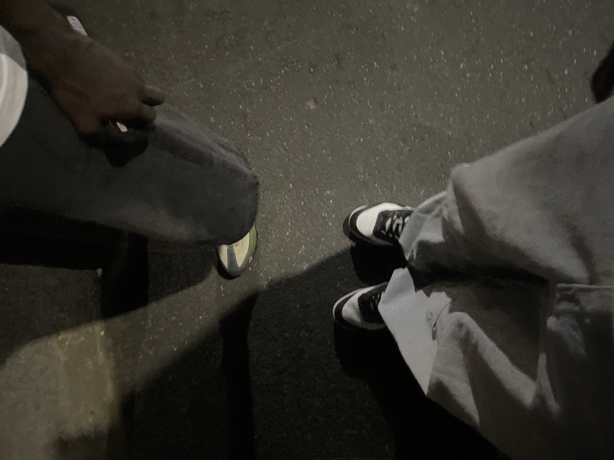 The image shows a view of two individuals standing on a paved surface, likely a street or sidewalk. The person on the left is wearing dark pants and a white shirt. A dark hand is visible. On the right, the individual is wearing light gray jeans and black and white sneakers. The environment appears to be urban, with dark asphalt possibly indicating nighttime. There is no information to indicate the location is Abuja, Nigeria.