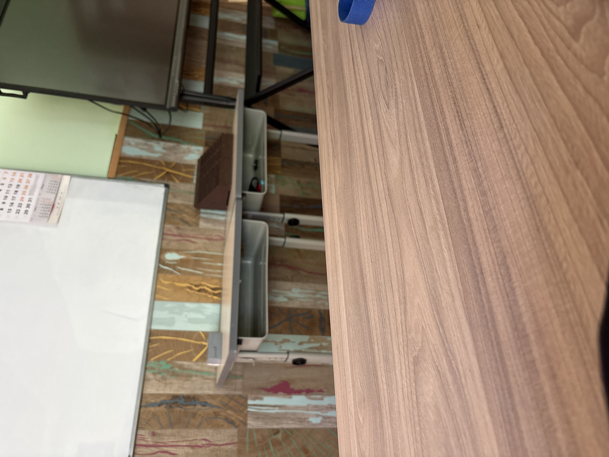 The image shows a classroom setting. In the foreground, a wooden desk surface is visible, with a blue strap in the lower right corner. In the midground, a pair of student desks with built-in compartments are arranged side-by-side against a wall with a decorative wood-paneled design featuring vertical stripes of various colors and wood grains. A brown object, possibly a stapler or organizer, sits on top of the right desk. Behind the desks, a large whiteboard occupies the left side of the wall, and a calendar is attached to it. To the right of the whiteboard is a light green wall section, and further to the right, a portion of a large screen or monitor on a stand is visible. The overall impression is that of an empty classroom in Redina, Bulgaria.