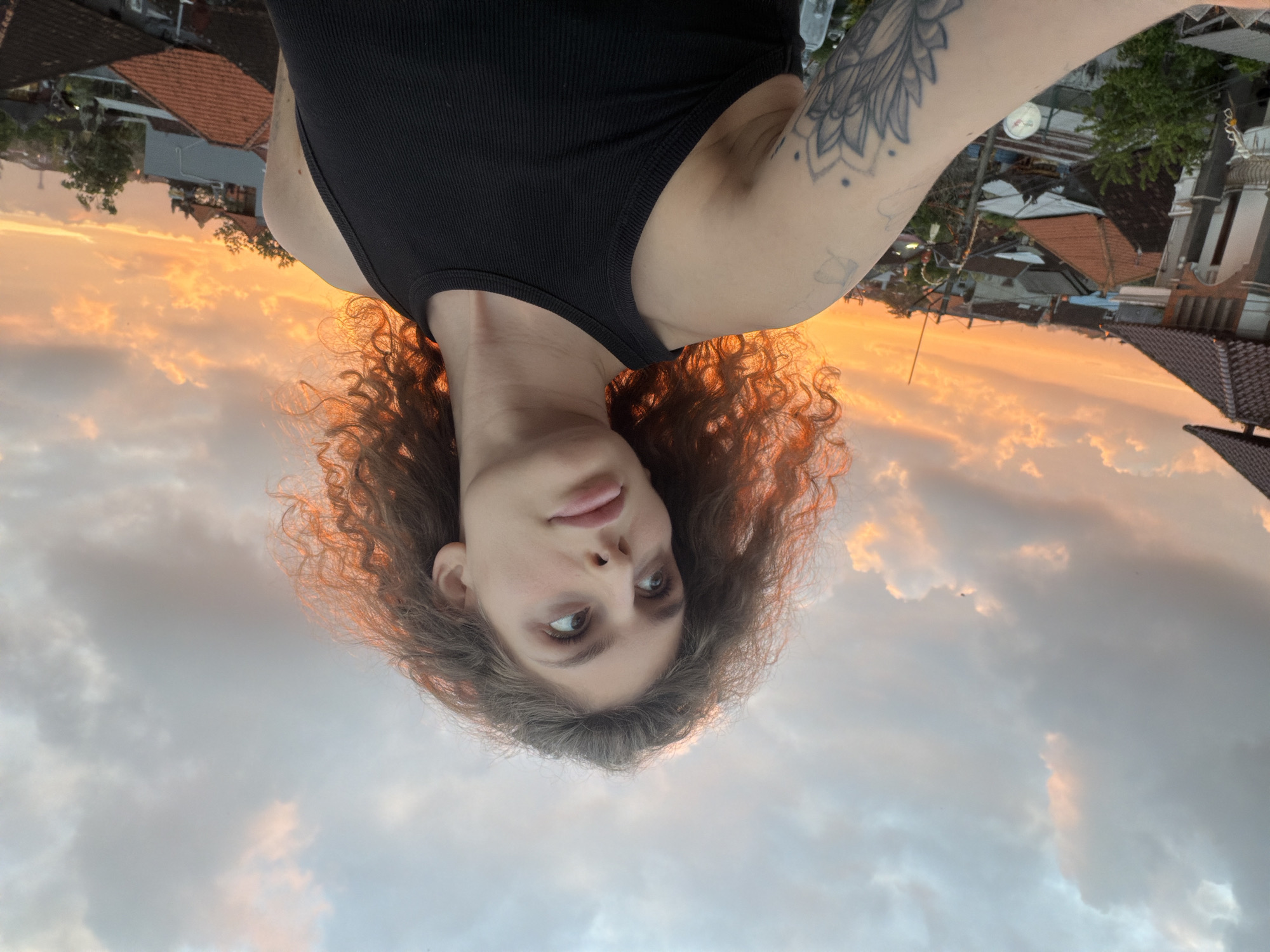 A medium shot captures a young woman with curly brown hair and fair skin posing for a selfie. She is wearing a black tank top and has a tattoo on her left arm. The background reveals a sunset over a city in North Kuta, Indonesia. The sky is filled with orange and pink clouds, illuminating the rooftops of houses and buildings. The woman is looking to her right, with her head tilted slightly upwards.