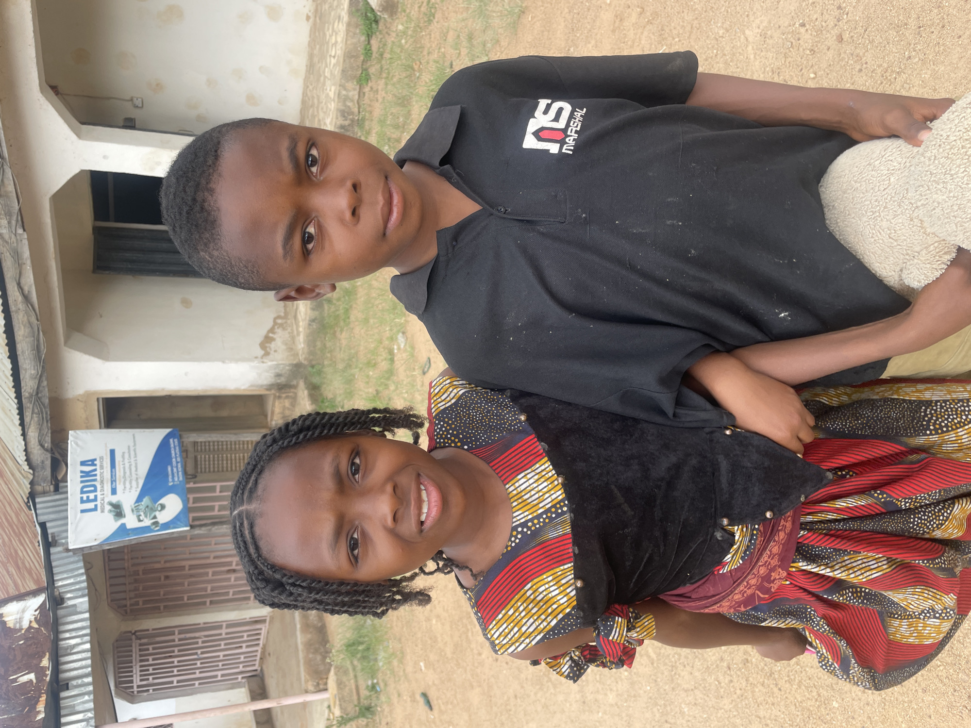 Two young individuals, a boy and a girl, stand close together outdoors, looking directly at the camera with smiles. The boy, positioned slightly behind the girl, has his right arm around her. He wears a black polo shirt with a white and red logo on the left chest that reads "MS MARSHAL." He holds a light-colored, textured object, possibly a small blanket or towel, in his left arm.
The girl, standing in front, has braided hair and is dressed in a vibrant, patterned dress. The upper part of her dress is black velvet with small metallic accents, while the skirt is adorned with colorful red, yellow, black, and white patterns, characteristic of African print fabric. Both children appear happy and friendly.
The setting is an outdoor area with a dirt or sandy ground. In the background, there are light-colored buildings with some signs of wear. To the left of the girl, a blue and white sign is visible, advertising "LEDİKA MEDICAL & DIAGNOSTIC SERVICES." The sign lists services such as "Blood Testing & Screening," "Urinalysis & Cultures," "X-ray Services," "Ultrasound Scans," and "Drug Test." It also displays the address "No. 40, Akawo Road, Barakin Akawo, Kaduna State, Nigeria." The scene appears to take place during the daytime in a community area in Barakin Akawo, Nigeria.