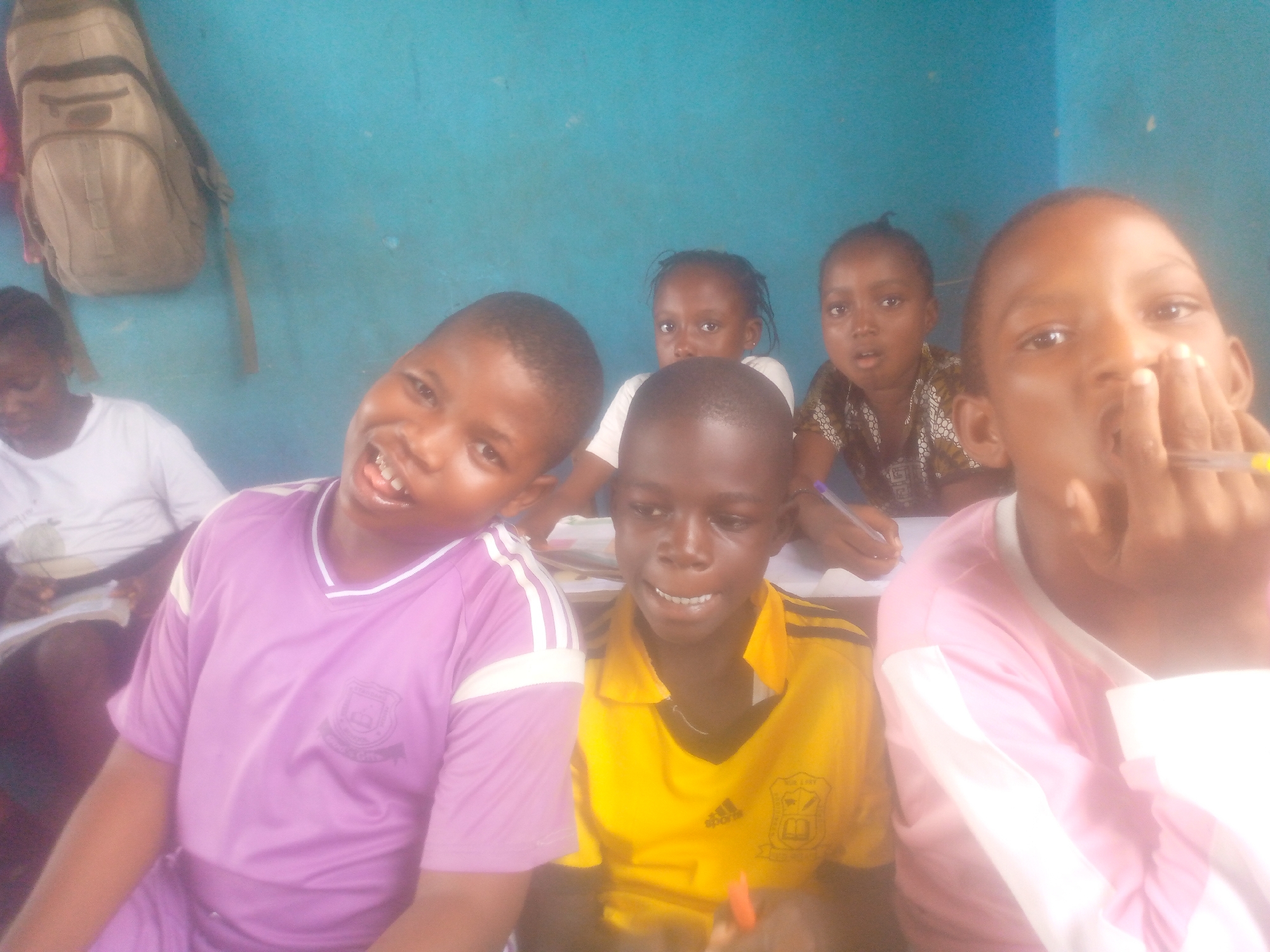 The image shows a group of students in a classroom setting. The location is in the city of Unknown, Unknown. The students are of African descent and appear to be young, possibly elementary school age. Some are seated at desks with paper in front of them, suggesting they are engaged in a writing activity. The environment is a simple classroom with a blue wall and a backpack hanging on it. The students' attire suggests they are wearing school uniforms.