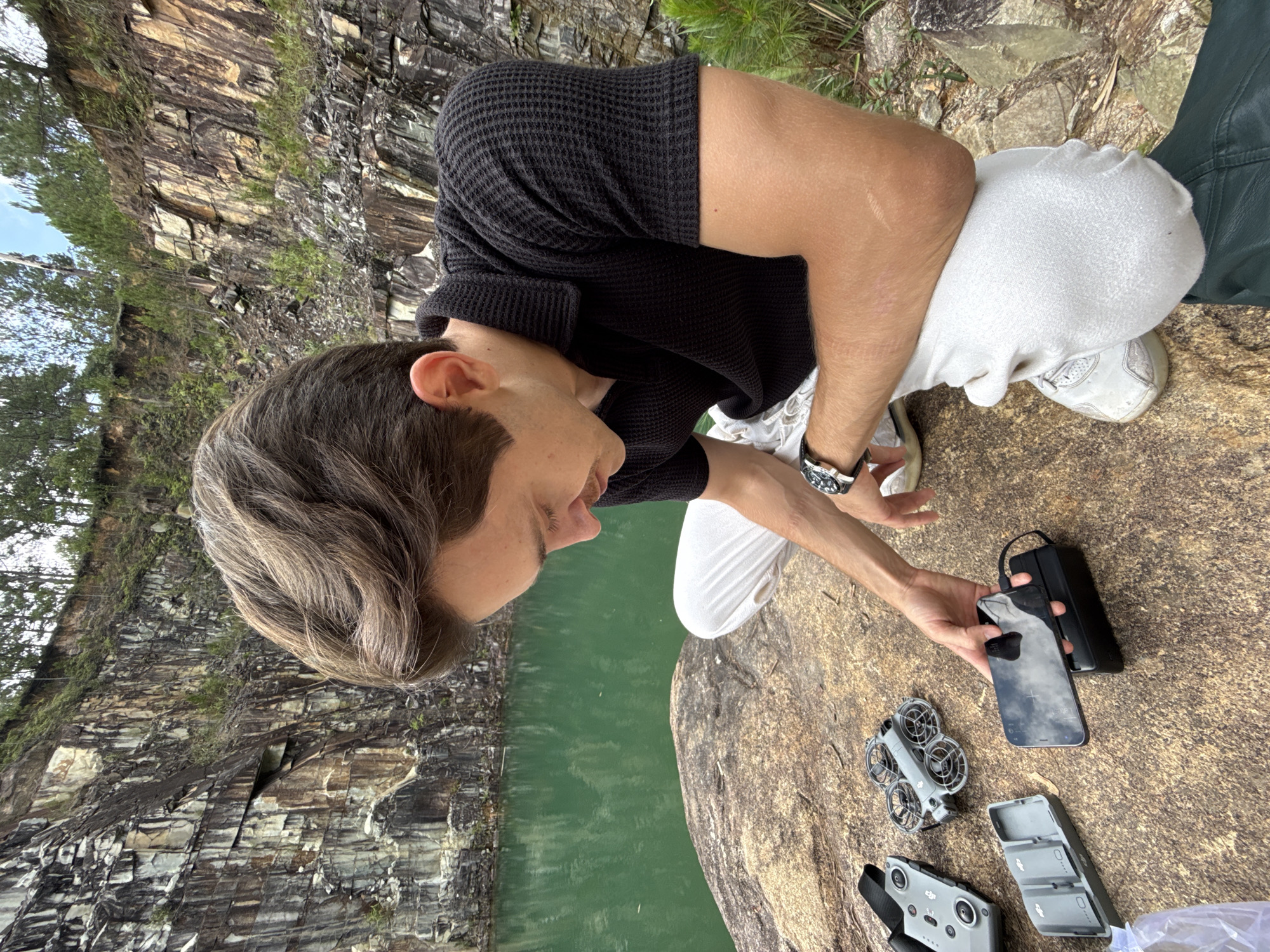 The image depicts a man kneeling on a large, rough rock formation next to a body of green water, likely a lake or quarry. The background shows a steep rock face with vegetation. The man is wearing a black short-sleeved knit shirt, white pants, and sneakers. He has a watch on his left wrist.
On the rock surface in front of him are several items: a smartphone connected to a power bank, a disassembled drone, a remote controller, and a multi-battery charging station. The man appears to be examining the phone or connecting it to the power bank.
The location is identified as Lang Biang - Da Lat, Vietnam based on the scenery.