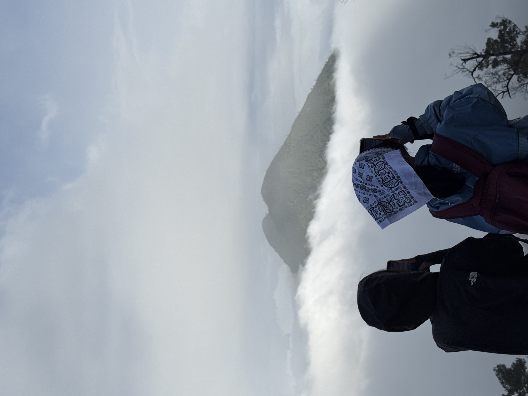 Two people are standing on a high vantage point, taking photos of a mountain peak shrouded in fog. The person on the left is wearing a black hooded jacket with "THE NORTH FACE" logo visible. The person on the right is wearing a blue jacket with a maroon backpack, their head covered with a white bandana. The sky is overcast with heavy clouds, and the landscape is dominated by mist and the grey mountain peak. The location is potentially in the city of Unknown, Indonesia, but no specific landmarks or details can confirm that from the photo.