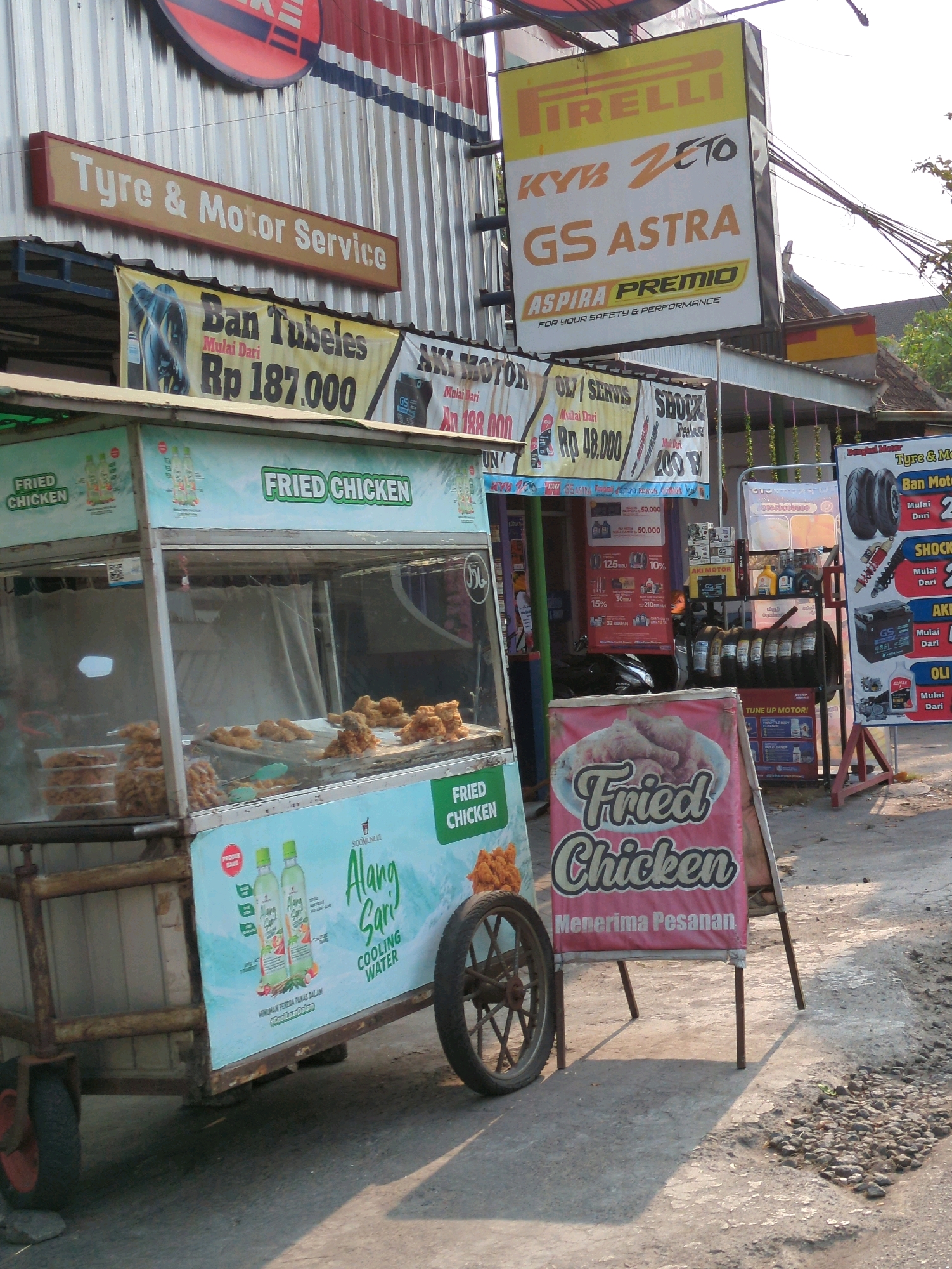 The image captures a street-level view of a commercial establishment in Blulukan, Indonesia, likely a roadside stall or small shop specializing in automotive services and food. The foreground features a mobile food cart, predominantly white and light blue, displaying "Fried Chicken" prominently on its side and front. The cart is laden with fried chicken displayed in a glass case, with additional fried items visible. Two green bottles of "Alang Sari Cooling Water" are advertised on the cart's side. A red wheel is attached to the cart's base. To the right of the food cart, a red A-frame sign advertises "Fried Chicken" with the text "Menerima Pesanan" (Accepting Orders).
Behind the food cart, the background reveals a building with a corrugated metal facade. Several signs are mounted on this facade. A prominent horizontal sign in beige and red reads "Tyre & Motor Service." Above this, a larger square sign displays the Pirelli logo, followed by "KYB ZETO" and "GS ASTRA," with "ASPIRA PREMIO" and "FOR YOUR SAFETY & PERFORMANCE" below. Further to the right, another sign with a motorcycle tire and shock absorber advertisement is visible. In the mid-ground, a rack is stocked with automotive parts, including tires, oil bottles, and spark plugs. The overall environment suggests a bustling street with various commercial activities. The lighting indicates it is daytime, with sunlight casting shadows on the ground.