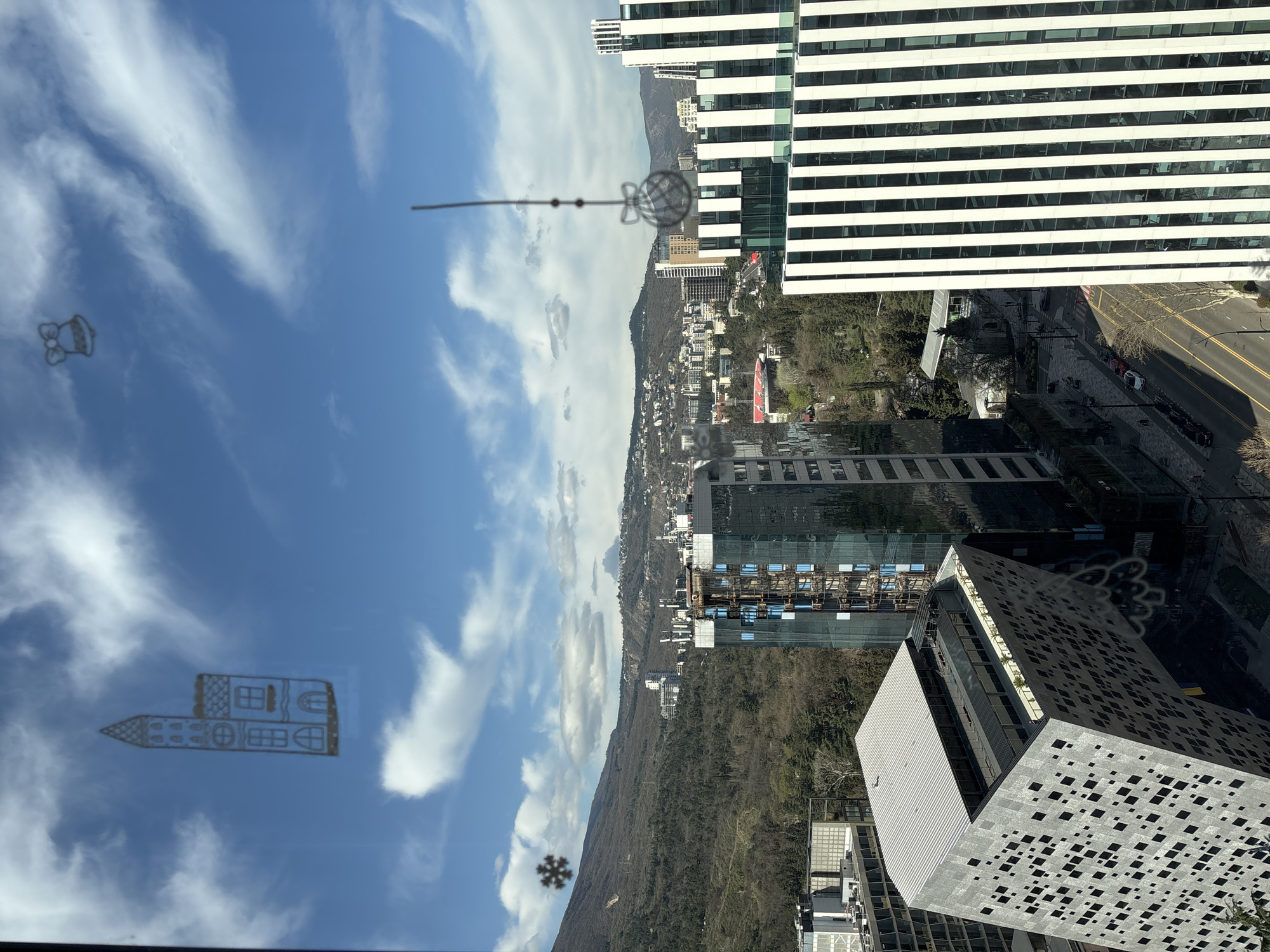 The image depicts a view from a high vantage point, possibly a window of a tall building, overlooking a cityscape in Tbilisi, Georgia. The sky is visible, with scattered white clouds against a blue background.
A large, modern building with a striped facade is partially visible on the right side of the frame. Below, other buildings are seen, including a structure with a perforated facade. A tree-lined street runs parallel to the buildings, with a few vehicles visible. A mountain range forms a backdrop to the cityscape.
Reflections are visible on the window, including a drawing of a house, a snowflake, a bell-shaped object, and other indistinct elements.