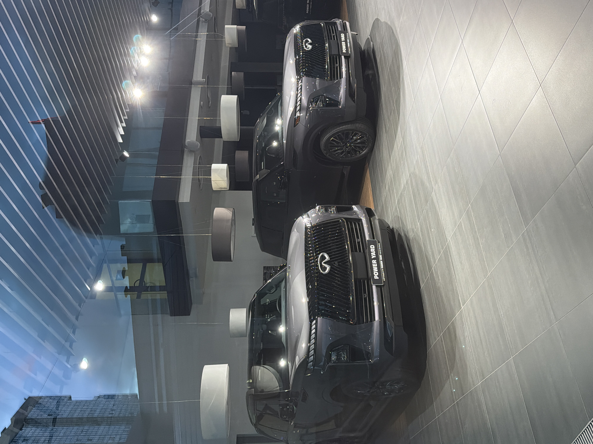 This is a photograph of the interior of a car dealership, likely in an urban environment, given the reflection of a tall building in the window. The image features two Infiniti QX80 SUVs, one in the foreground and another slightly behind it. Both vehicles are dark gray and parked on a tiled floor. The dealership's branding, "POWER YARD," is visible on a plate on the front of the foreground vehicle. Above the cars, cylindrical lamps hang from the ceiling. The background shows a mezzanine level with glass railings and more lighting. The overall impression is a modern and well-lit showroom.