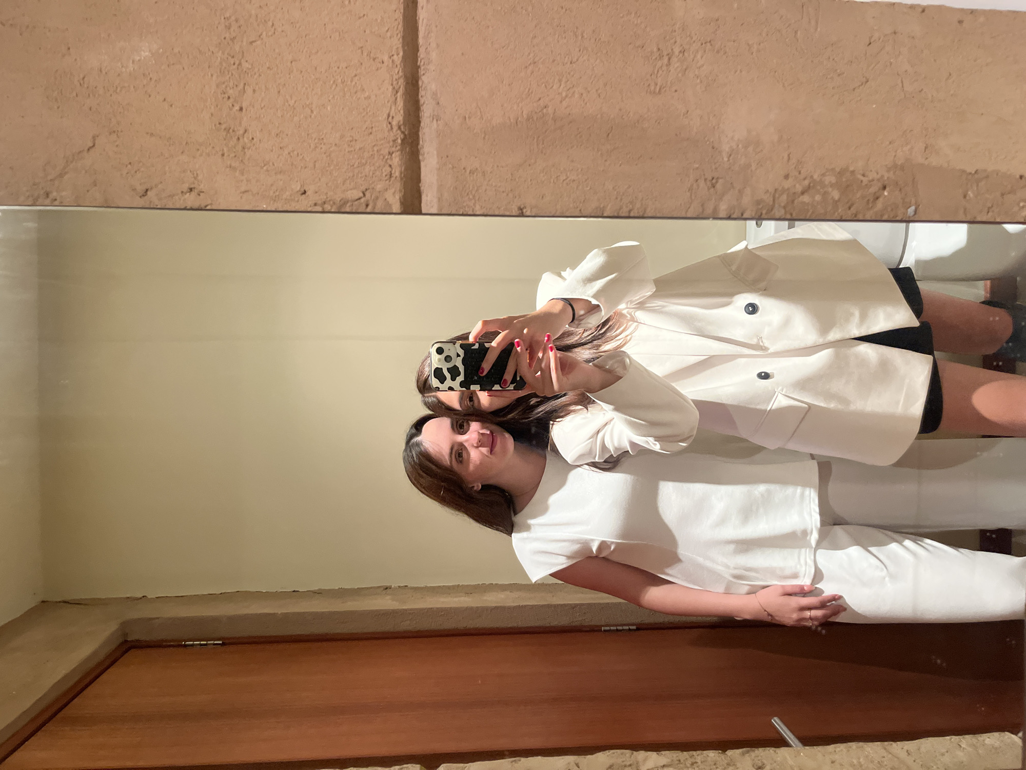 Two young women are reflected in a mirror, taking a selfie. The woman in the foreground wears a white t-shirt and white pants. The woman in the background wears a white blazer over a black top. The background appears to be an interior space with beige walls and a wooden door frame. The setting is in San Bernardino Tlaxcalancingo, Mexico.