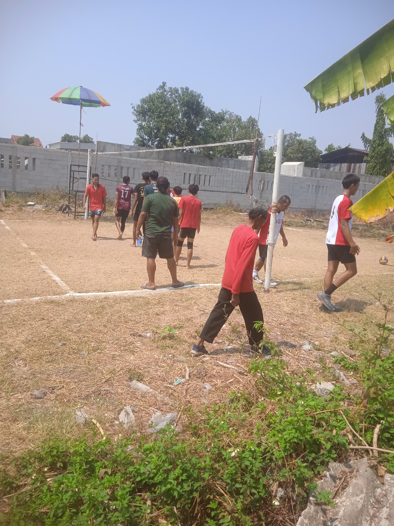In Klodran, Indonesia, a group of young men are gathered on a dry, grassy volleyball court under a clear sky. A volleyball net is stretched across the court, with poles supporting it. Several players are visible in motion, some wearing athletic attire. One individual in a red shirt and black pants is in the foreground, walking. Behind him, a player in a maroon jersey with the number 17 on it is also walking. In the background, a colorful umbrella is propped up on a wooden pole. The surrounding environment consists of a low brick wall, trees, and some scattered debris on the ground.