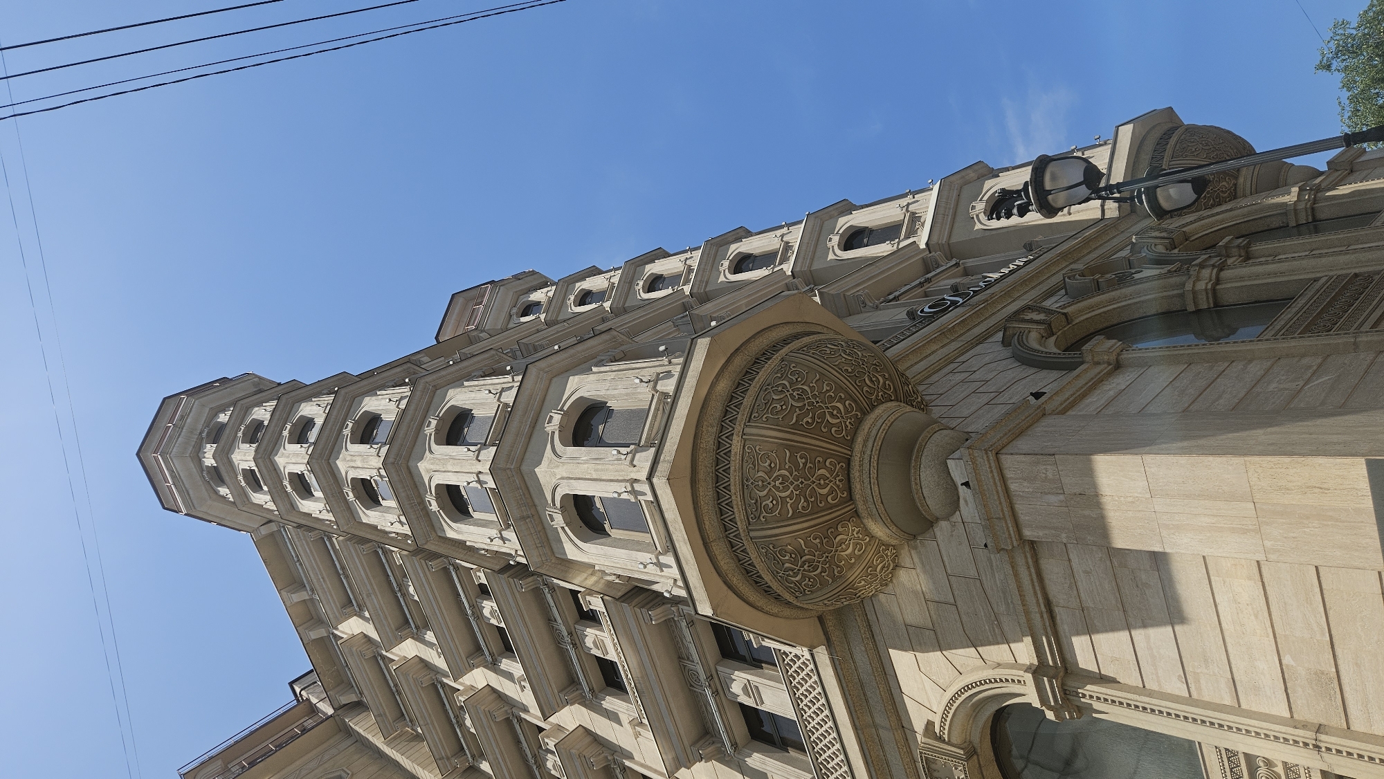 The image is a low-angle, upward shot of a detailed, ornate building corner in Almaty, Kazakhstan. The building is constructed of light-colored stone and features multiple floors with arched windows and decorative balconies. A prominent circular balcony with intricate carved detailing juts out from the corner. The building's corner structure extends upwards in a polygonal turret shape. Electrical wires are visible against a clear blue sky. A vintage-style street lamp with two lights stands near the base of the building. No people are visible in the image. The architectural style suggests a historical or classic design.
