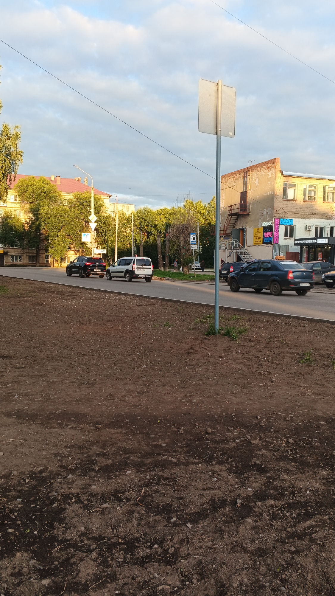 The image depicts a street scene in Izhevsk, Russia, likely on a cloudy day with soft lighting. In the foreground, a dirt patch with sparse vegetation and debris lines the edge of a paved road. Several cars are visible on the road; a black SUV and a white minivan are in the left lane, while a dark sedan is in the right lane, followed by other vehicles. The road is bordered by trees and buildings. On the left side, a multi-story building with a red roof and trees in front of it can be seen. On the right side, a weathered, brick building with an external staircase and several windows stands. A prominent street sign, a white rectangular placard on a pole, is positioned in the center of the image. The sky above is partially covered with clouds.