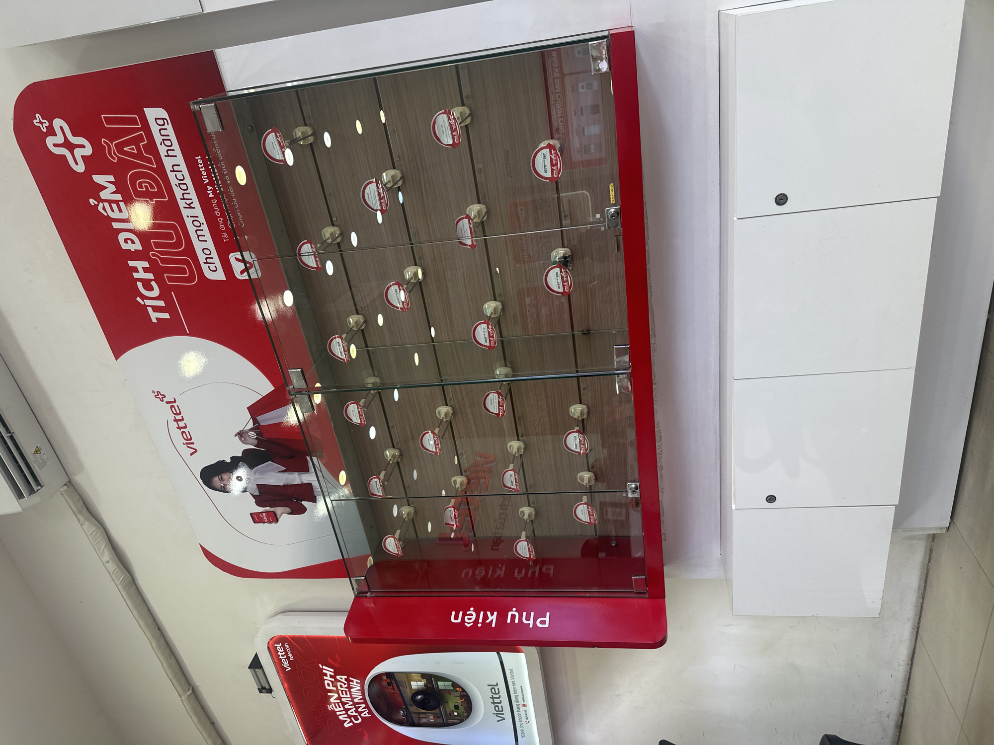 The image captures an indoor scene, likely a retail store or service center, characterized by its modern and organized appearance. The primary focus is a large, vertical red and white display stand situated in the center-left. This stand features a prominent glass-fronted cabinet.
On the main red panel of the display, large white text reads "TÍCH ĐIỂM ƯU ĐÃI" (Earn points for offers) and "cho mọi khách hàng" (for all customers). Below this, smaller text instructs "Tải ứng dụng My Viettel" (Download My Viettel app). A graphic of a woman in red holding shopping bags and a smartphone, accompanied by the Viettel logo, is also part of this display.
Inside the glass cabinet, numerous small, white hooks are arranged on shelves, each holding a red-and-white tag. Many of these tags feature the Viettel logo and the text "GIÁ TỐT" (Good Price), along with partially obscured phone numbers like "0979***88%" and "0378***567". The reflection on the glass shows mirrored Vietnamese text, including "Chọn ưu đãi và đổi điểm dễ" (Choose offers and redeem points easily). Below the glass section, a red panel has the word "Phụ kiện" (Accessories) written in white.
To the immediate left of the main display, a smaller red and white stand showcases a security camera. Text on this stand includes "SHOCK", "MIỄN PHÍ CAMERA AN NINH" (Free Security Camera), and "Dành cho khách hàng dùng Internet Viettel" (For customers using Viettel Internet). An air conditioning unit is visible further left.
On the right side of the image, a stack of white, lockable cabinets or drawers stands against a plain wall. The overall setting is brightly lit by artificial light, indicating an indoor commercial environment. No specific activity or event is taking place; the scene is static, presenting product displays and promotional information. The presence of Viettel branding and Vietnamese text confirms the location in Vietnam, specifically in Nha Trang Ward as per the provided context.