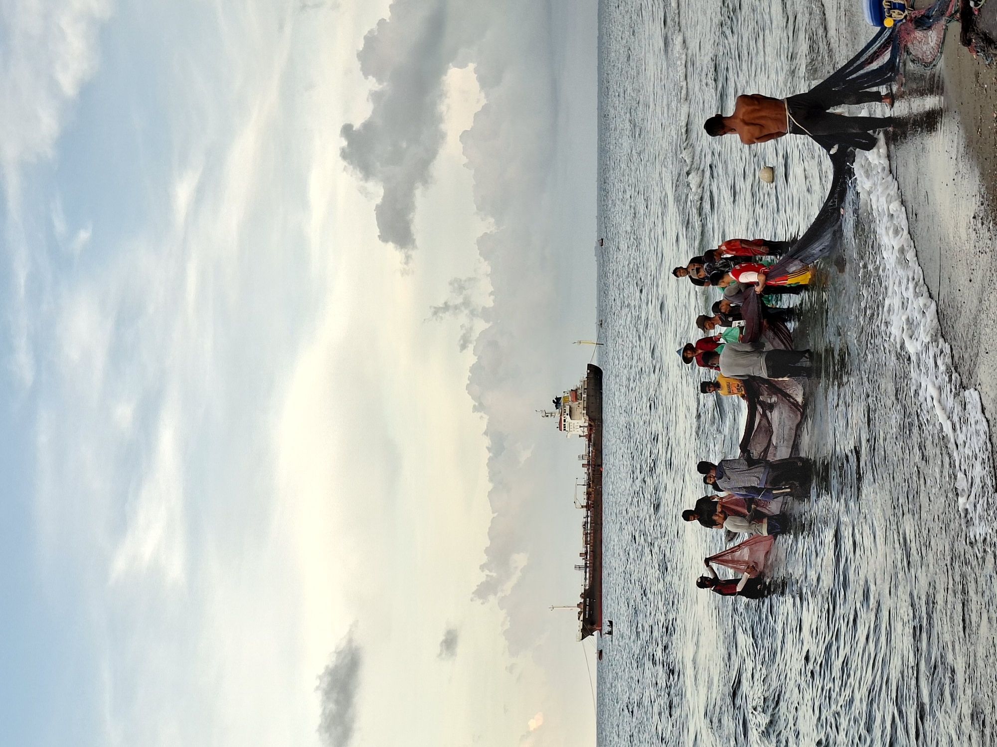 A slightly rotated medium shot depicts a group of people fishing along the shoreline in Lhokseumawe, Indonesia. The water is choppy, and a large ship is visible in the distance, obscured by clouds in the sky. The group is pulling a large fishing net from the water. One shirtless man is pulling the net from the shore. The overall lighting is soft, suggesting the photo was taken during sunrise or sunset. The scene conveys a sense of community and local tradition related to fishing.