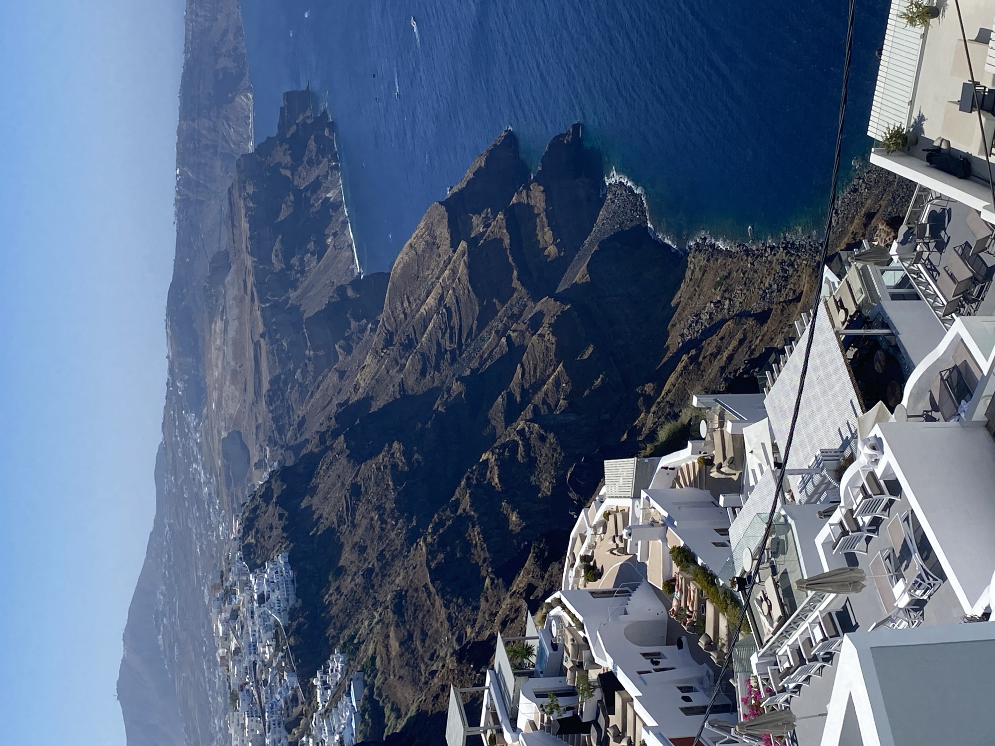 This image captures a panoramic view of the caldera in Santorini, a Greek island in the Aegean Sea. In the foreground, white, multi-level buildings with flat roofs, characteristic of Cycladic architecture, cascade down the cliffside. These structures appear to be part of a resort or hotel complex, with balconies, terraces, outdoor seating areas, lounge chairs, and umbrellas visible. The buildings are stark white, contrasting with the deep blue of the sea.
In the middle ground, the rugged, volcanic cliffs of the caldera rise steeply from the water. The terrain is predominantly barren, with dark, rocky slopes and sparse vegetation. Small white structures, likely part of the town of Thira, are scattered across the distant hills and clifftops, blending into the landscape.
The background features the vast expanse of the Aegean Sea, a deep, rich blue under a clear sky. A few small boats are visible on the water, and the distant horizon shows the continuation of the island's dramatic coastline. The overall impression is one of a breathtaking Mediterranean landscape, combining human habitation with the raw beauty of nature. The scene is set during daytime, under bright sunlight, creating sharp shadows and highlighting the textures of the rocks and buildings.