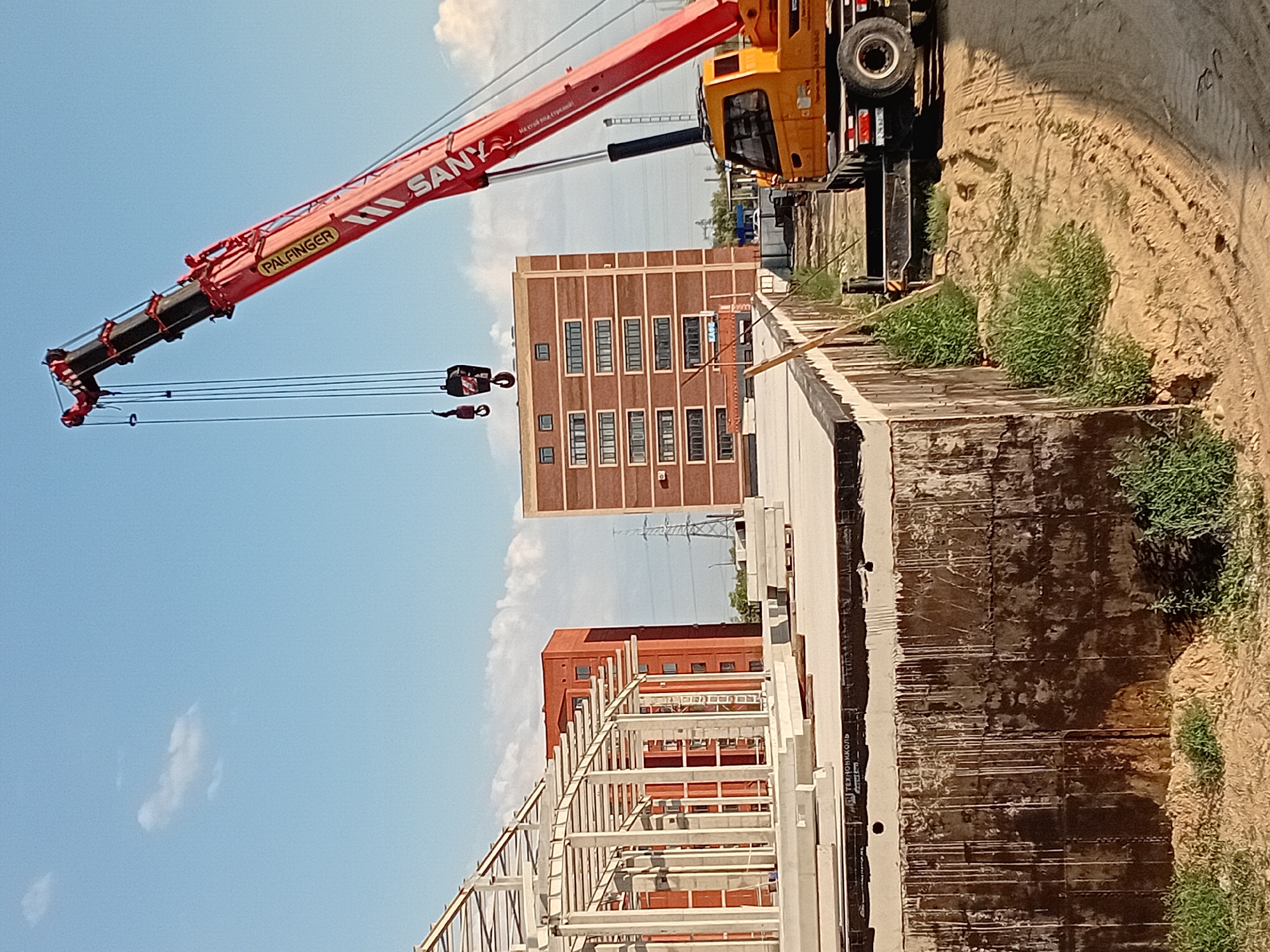 This image captures an outdoor construction site in the city of Semey, Kazakhstan, under a clear blue sky with sparse clouds. A large, red mobile crane, identified by "PALFINGER" and "SANY" branding, dominates the right side of the frame, its boom extended upwards. Below the crane, a yellow truck cab is visible, suggesting the crane is mounted on a truck chassis.
In the foreground, a partially constructed building with a concrete foundation and steel framework is visible. The foundation walls are dark and appear wet or stained. The framework consists of vertical concrete columns and horizontal steel beams, indicating the early stages of building construction.
Further in the background, two multi-story brick buildings stand. The one to the left is a darker shade of red, while the one to the right is a lighter red-orange. Both buildings have rows of windows. A tall, lattice-style electricity pylon can be seen between these buildings and the construction site.
The ground surrounding the construction area is unpaved, with visible tire tracks in the sandy soil, typical of a construction environment. Green vegetation, including grass and weeds, grows along the edge of the construction site and near the buildings. The overall scene depicts an active construction zone in a semi-urban or industrial area.